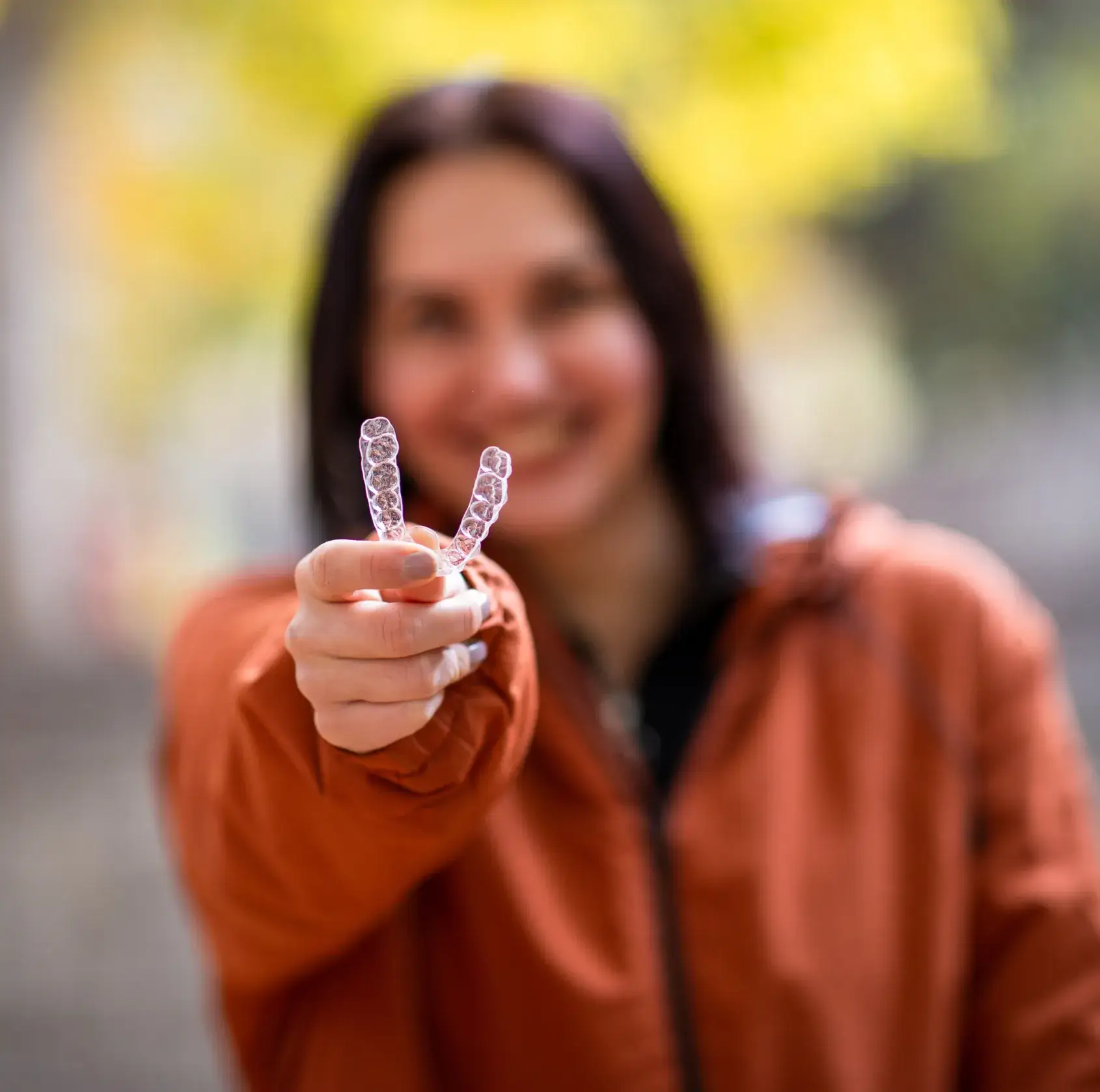 Woman in an orange jacket holding clear dental aligners towards the camera with a blurred green and yellow background.