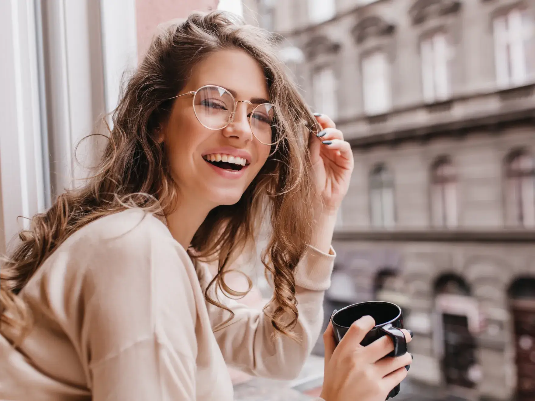 Smiling woman with curly hair wearing glasses holding a black mug near a window with a blurred city street background.