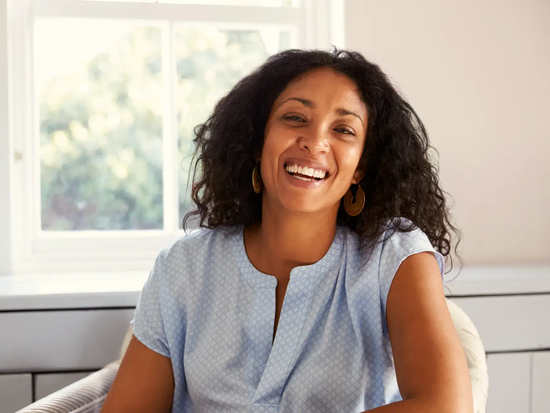 Smiling woman with curly hair wearing gold earrings and a light blue patterned blouse, sitting indoors near a window.