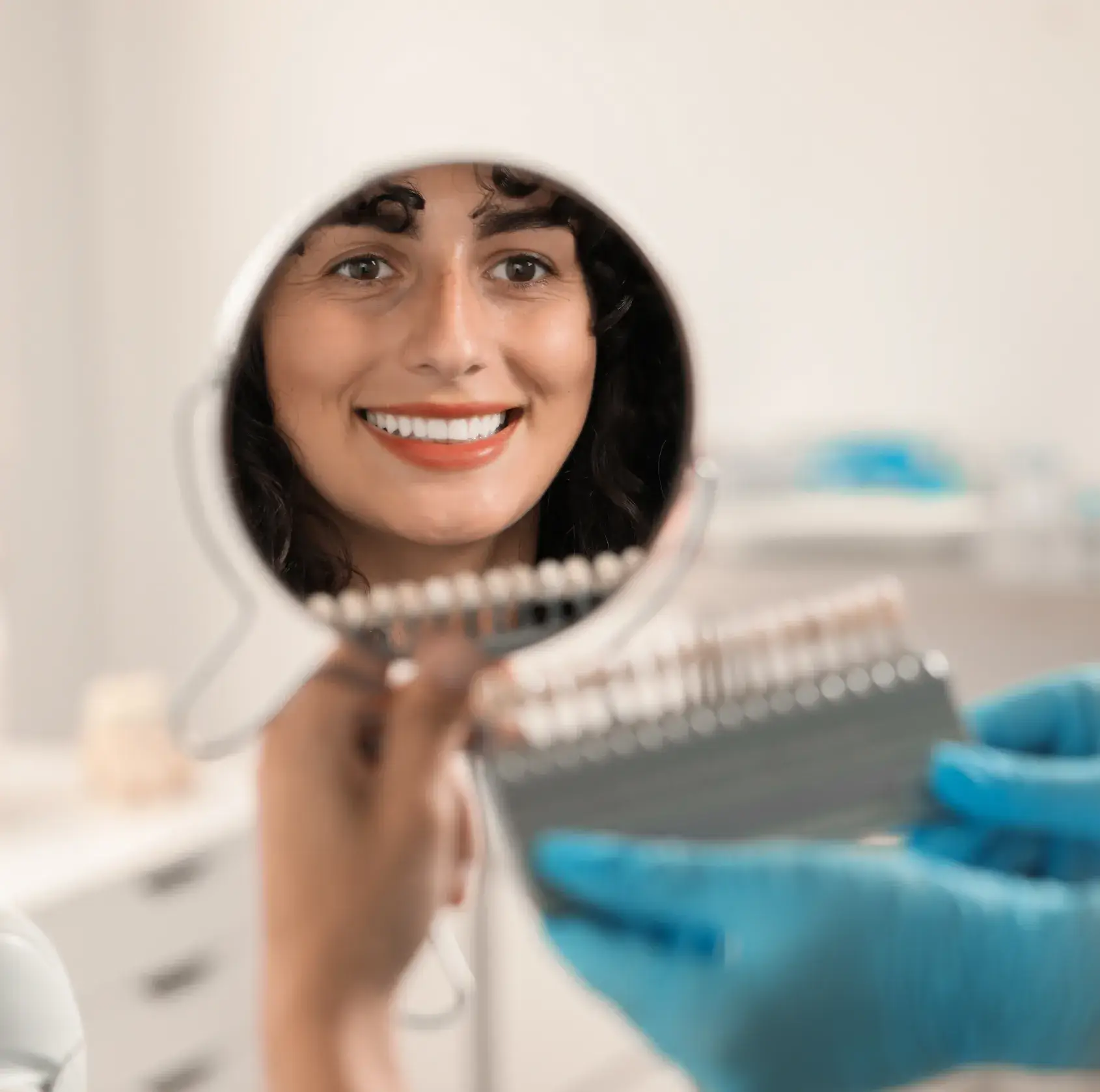 Woman smiling into a handheld mirror while a gloved hand holds a dental shade guide.