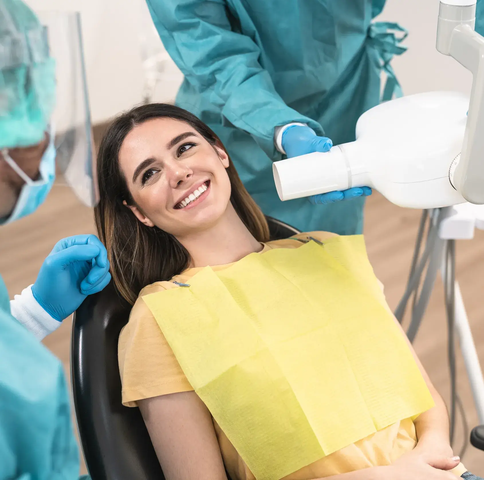 Smiling woman in a dental chair wearing a yellow bib while a dentist in protective gear prepares equipment.