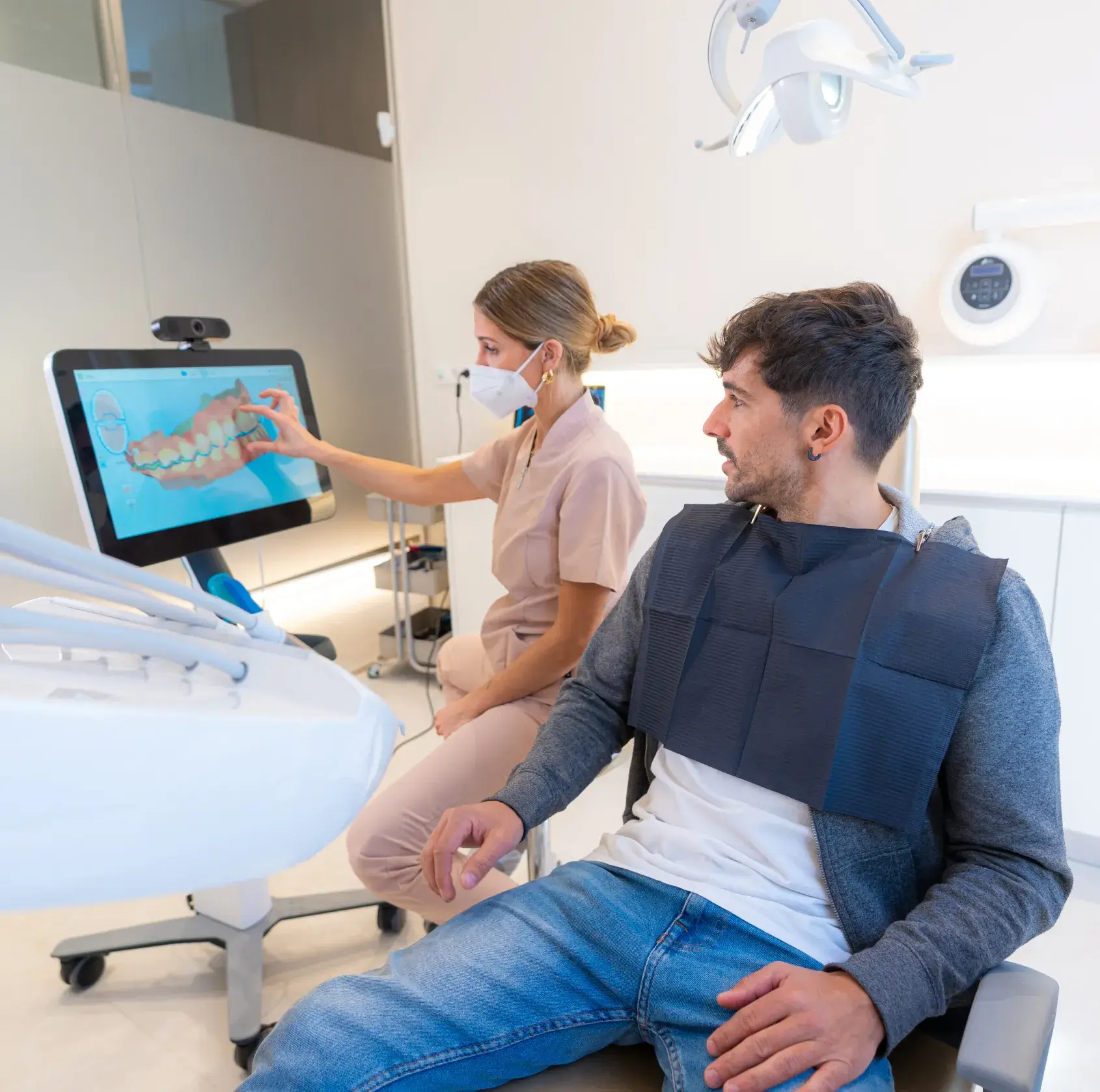 Dentist in scrubs and mask explaining digital dental X-ray to male patient sitting in dental chair.