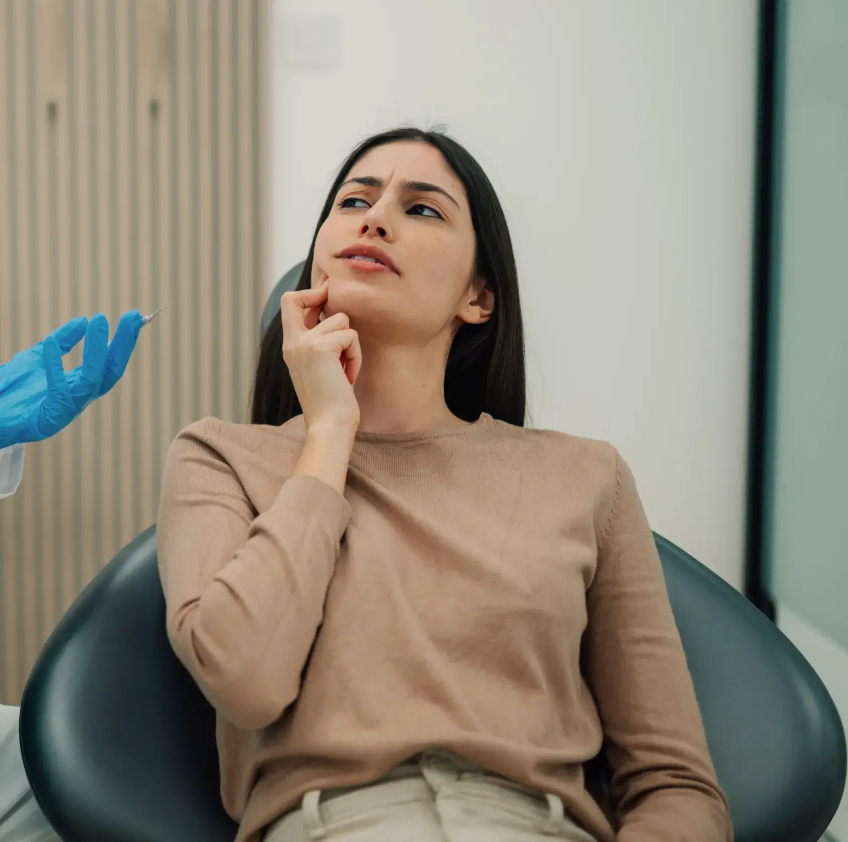 Woman sitting in a dental chair, touching her cheek with a concerned expression while a gloved hand holds a syringe nearby.