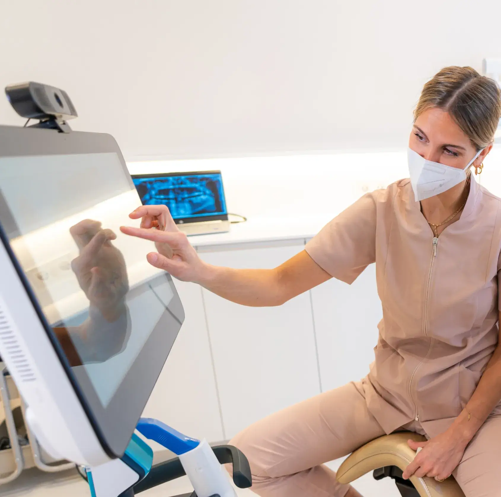 Dentist wearing a mask and scrubs interacting with a touchscreen monitor in a clinical setting with a dental X-ray on a laptop in the background.