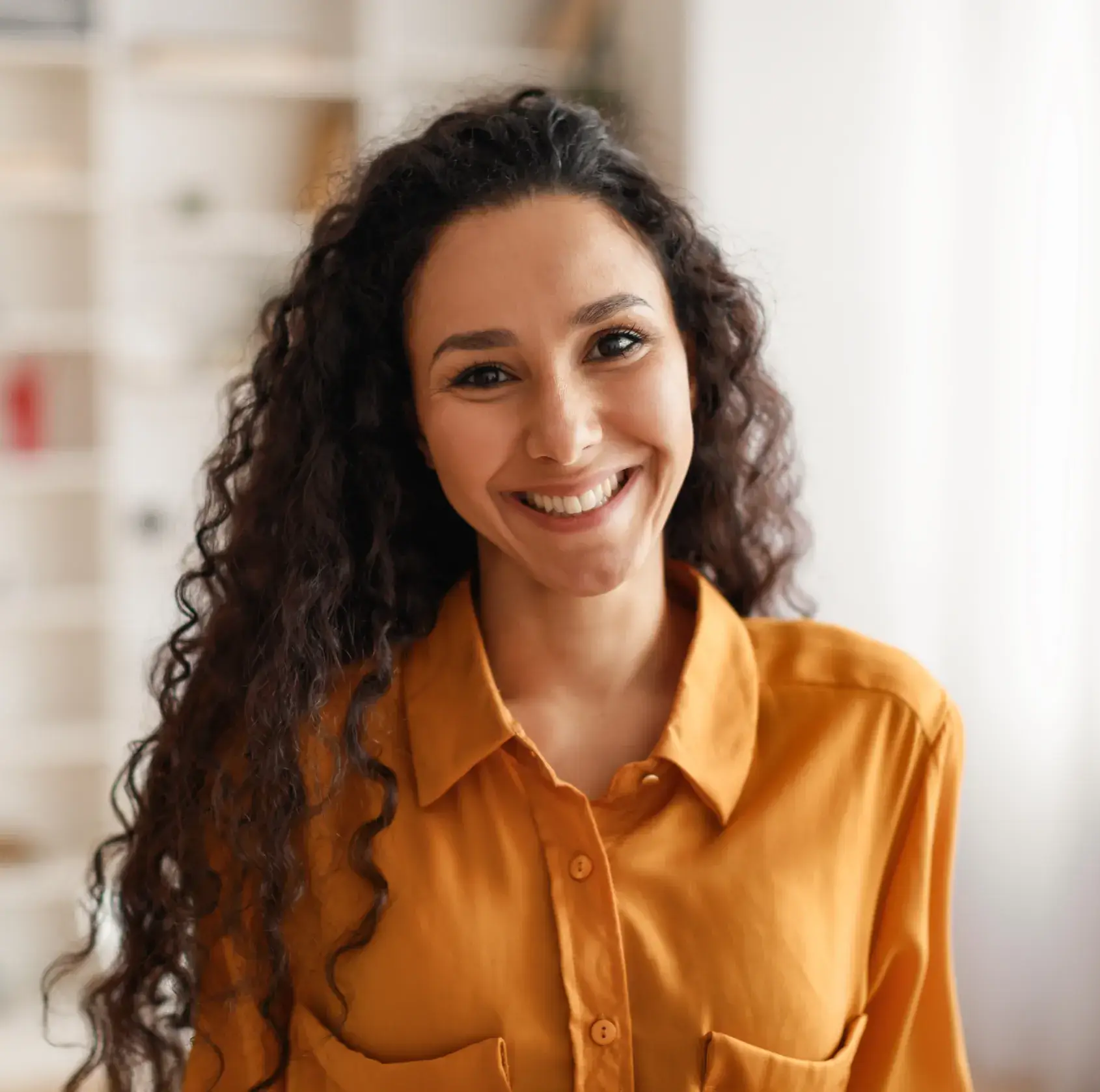 Smiling woman with long curly hair wearing a mustard yellow button-up shirt.
