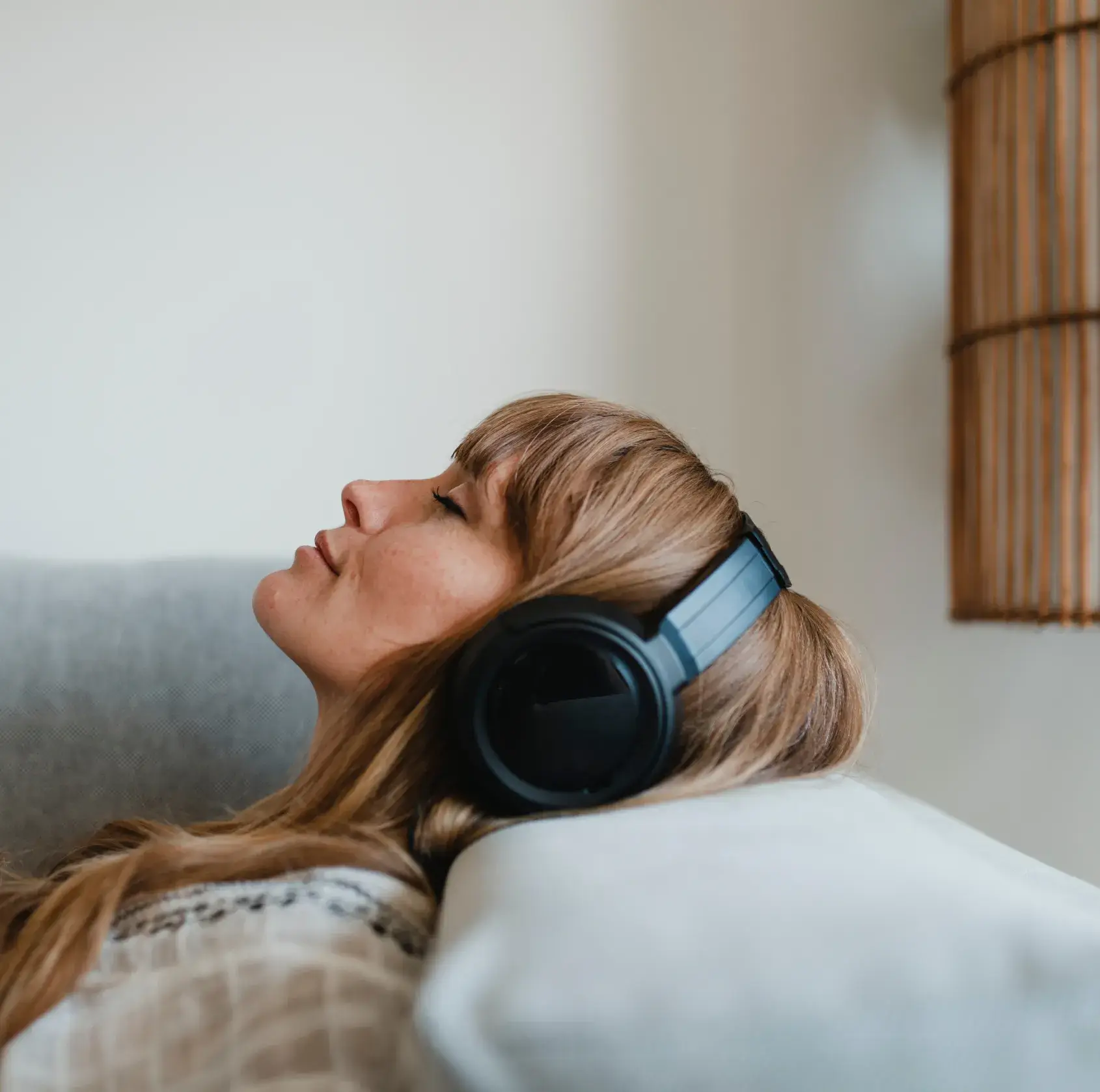 Woman with long blonde hair wearing black headphones and reclining on a gray couch with closed eyes.