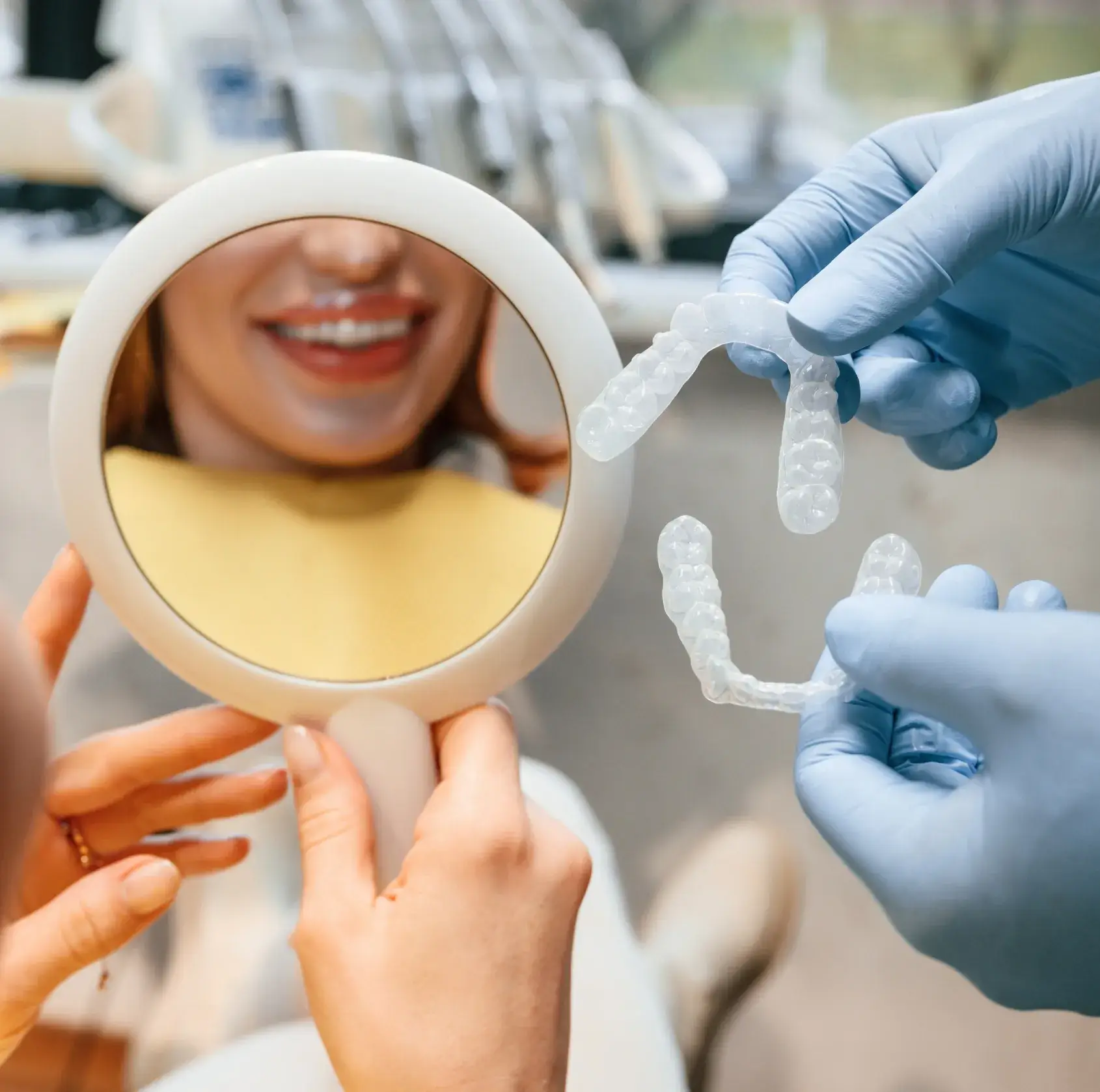 Patient holding a round mirror smiling at teeth while a gloved hand holds clear dental aligners.