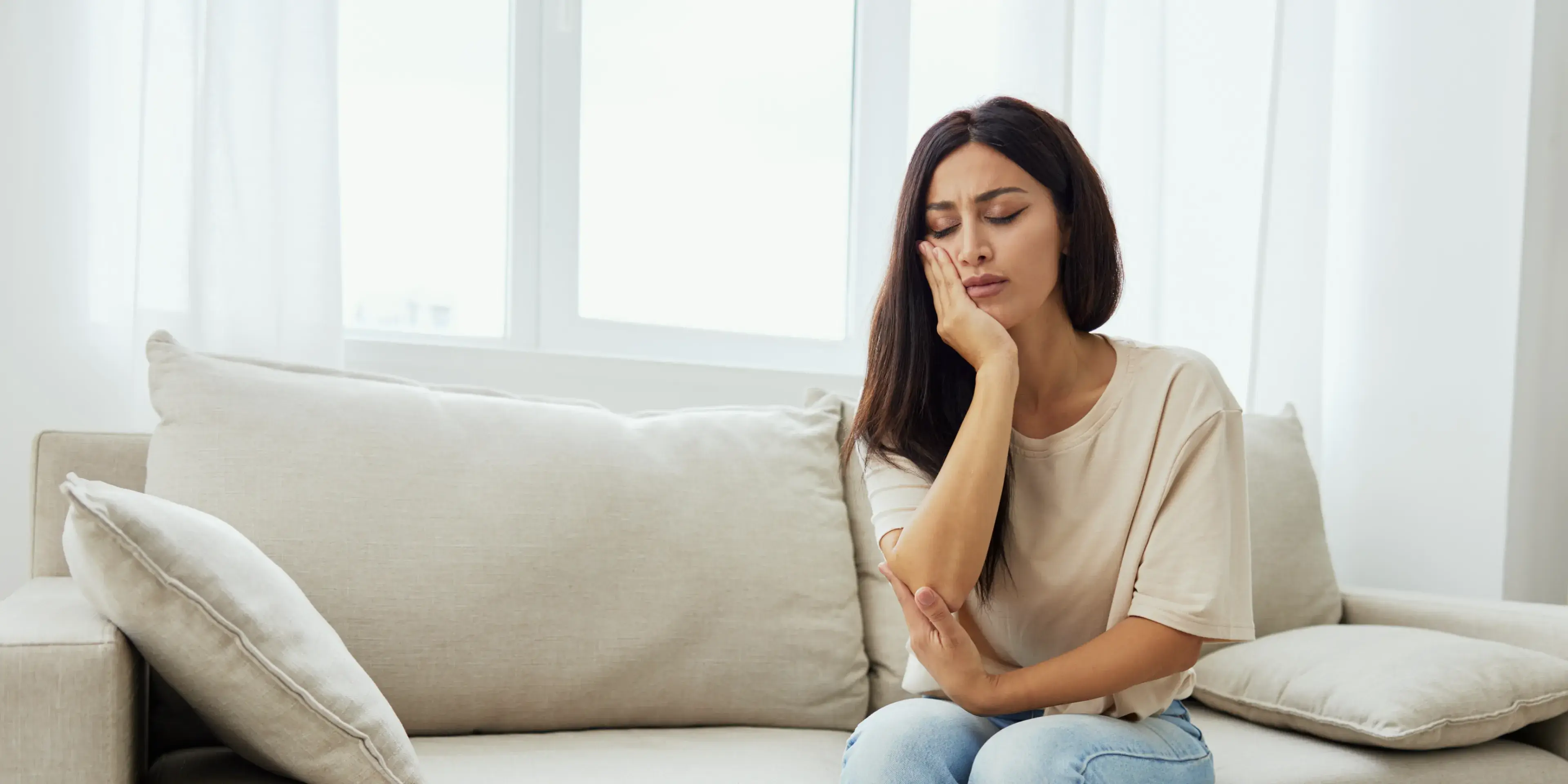 Woman sitting on a beige couch holding her cheek with a pained expression, indicating tooth or jaw pain.
