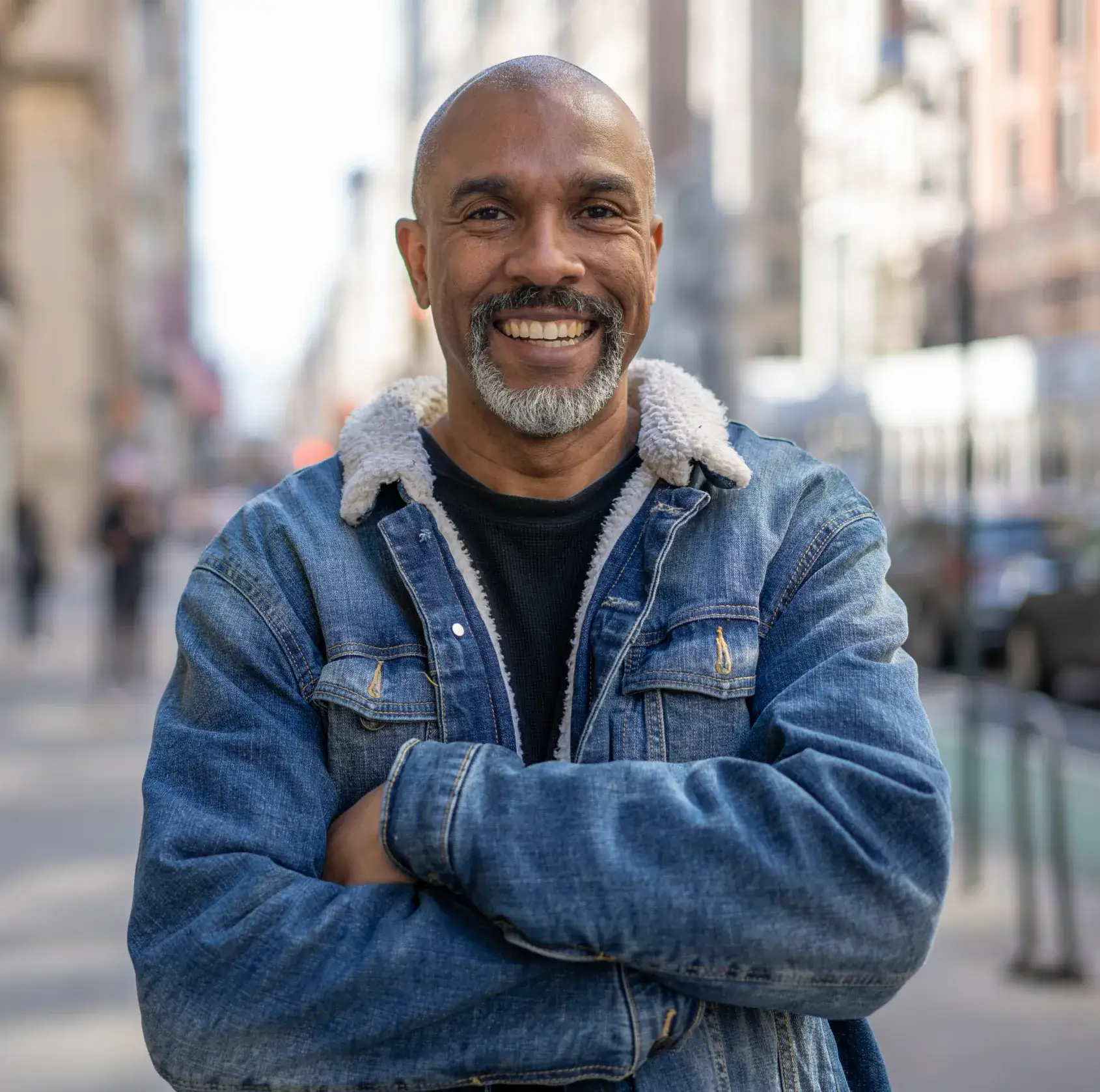 Smiling middle-aged man with gray beard wearing a denim jacket with fleece collar, standing with arms crossed on a city street.