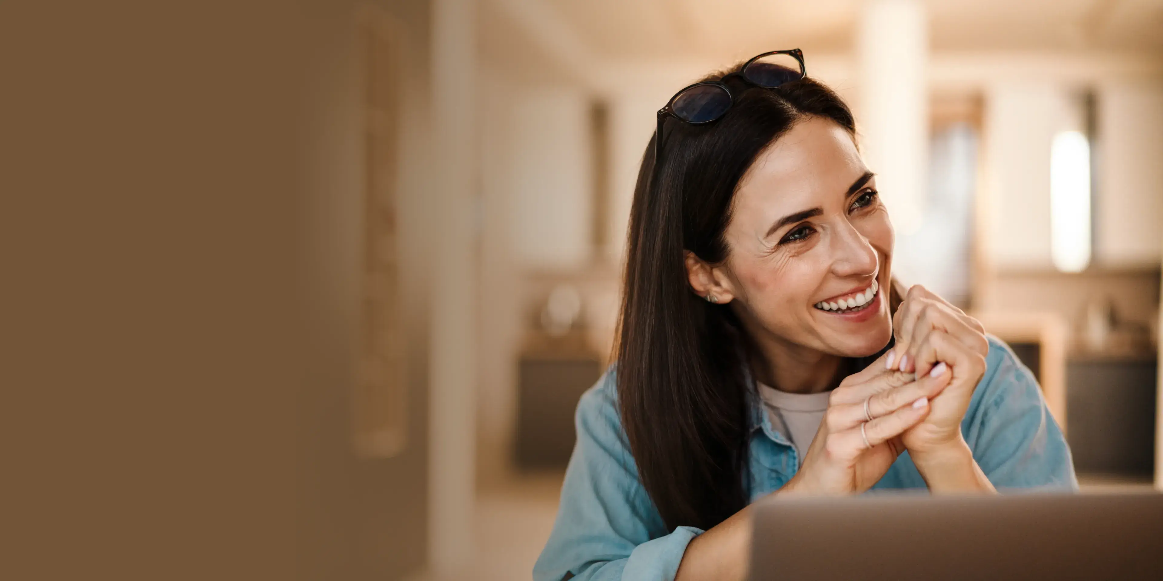 Smiling woman with dark hair and sunglasses on her head sitting indoors near a laptop.