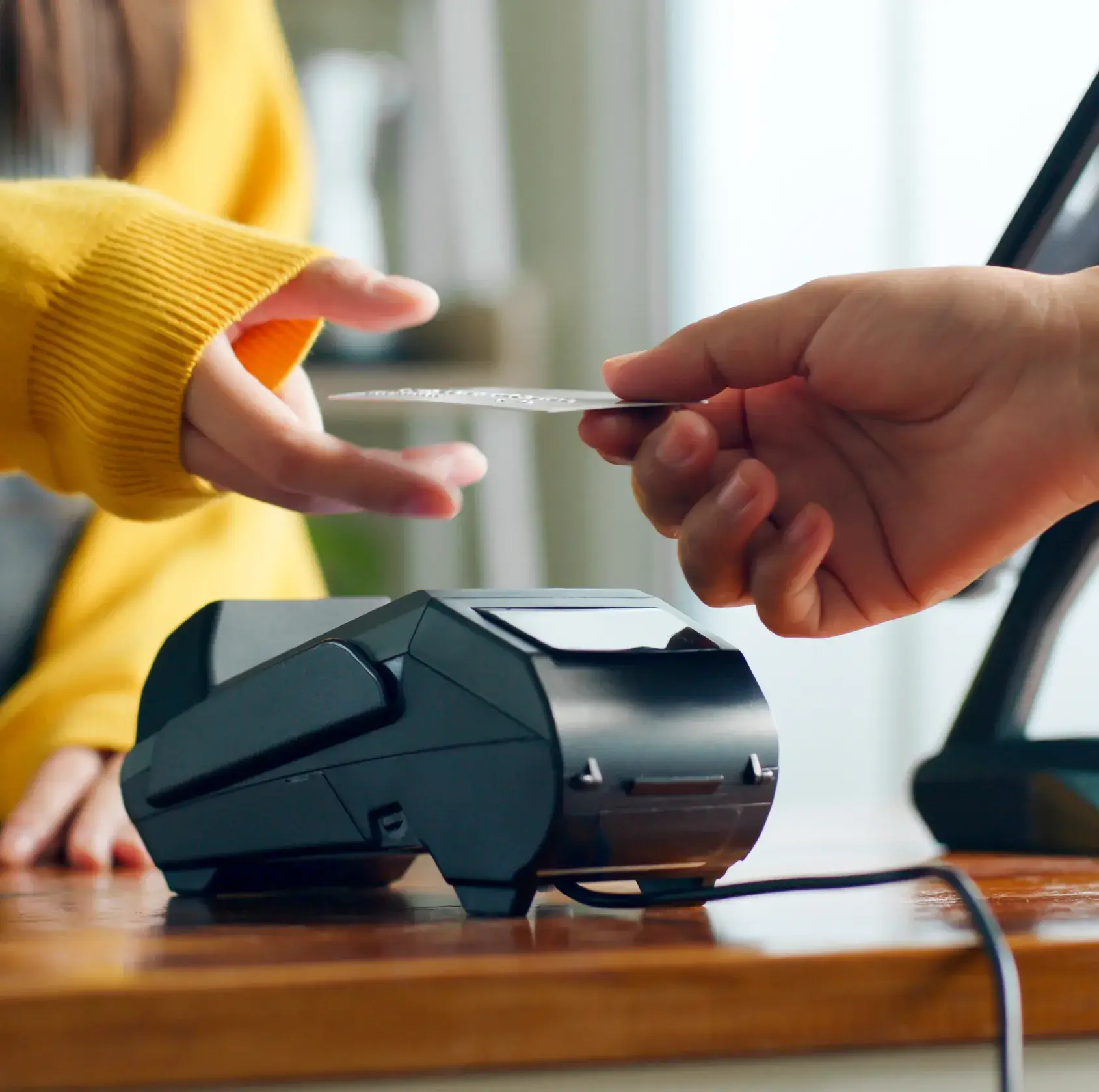 Person in yellow sweater reaching to take a card from another hand over a payment terminal on a wooden counter.