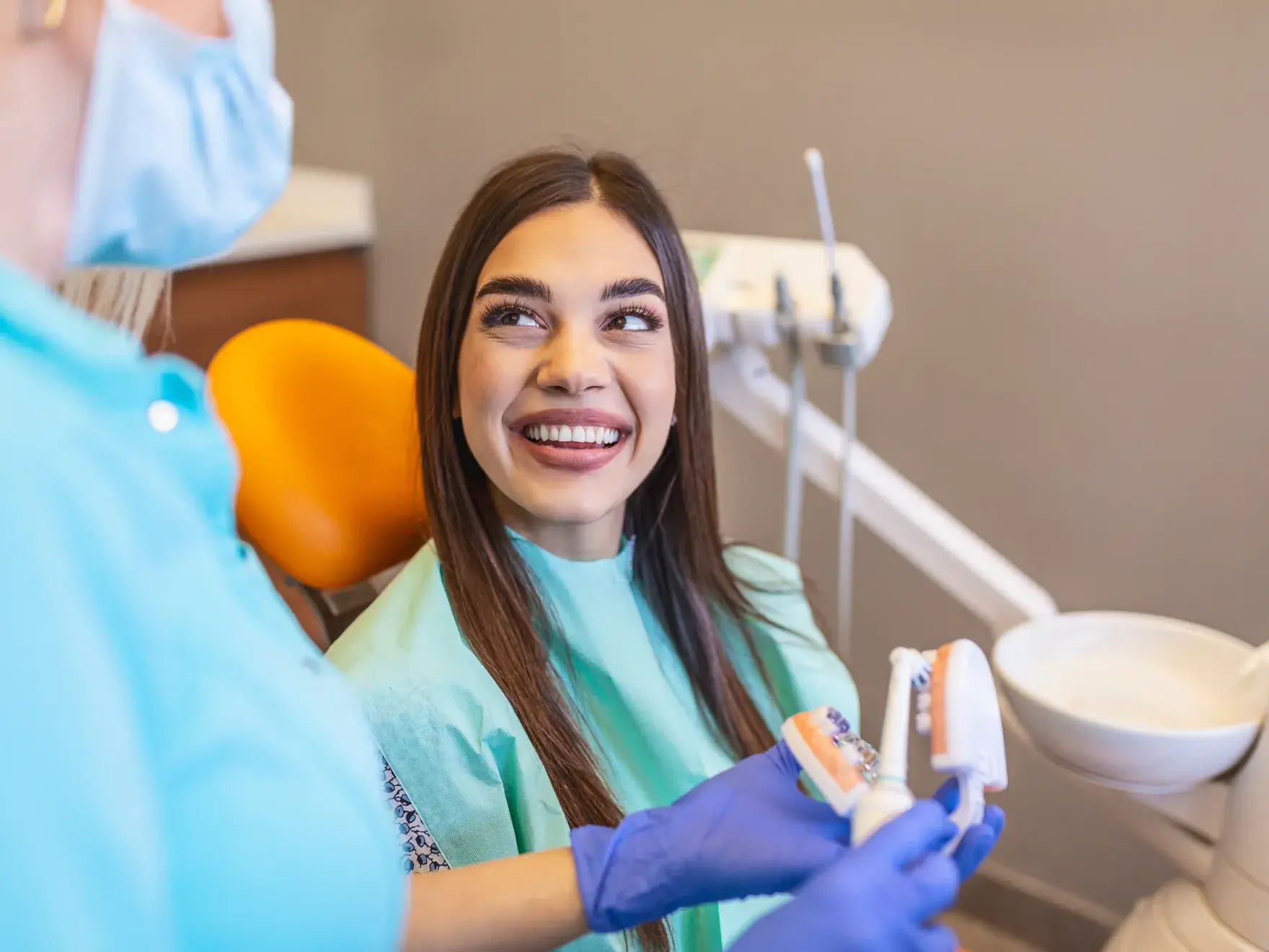 Smiling woman in a dental chair looking at a masked dental professional holding a dental model and toothbrush.