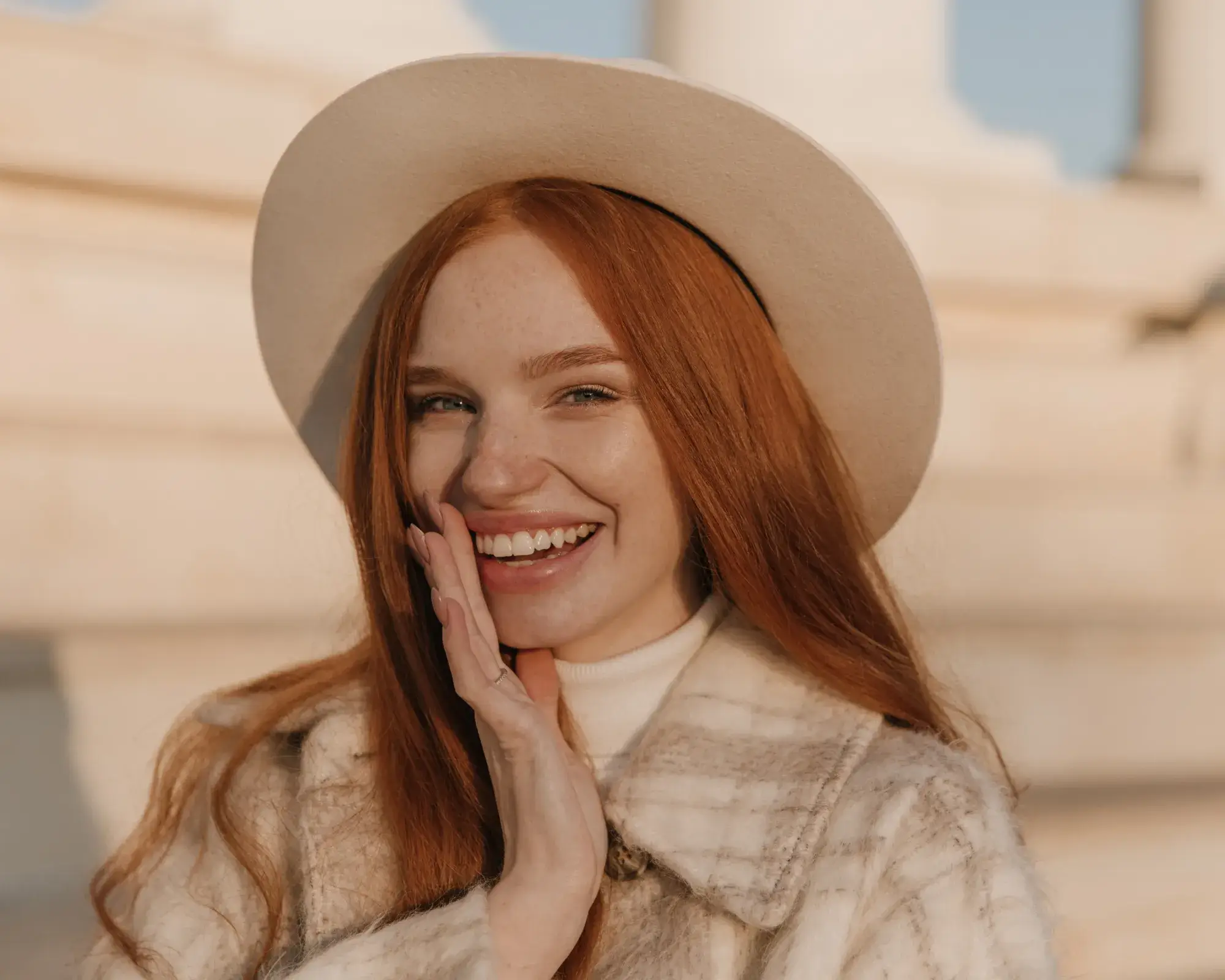Smiling red-haired woman wearing a beige hat and light plaid coat, touching her face.
