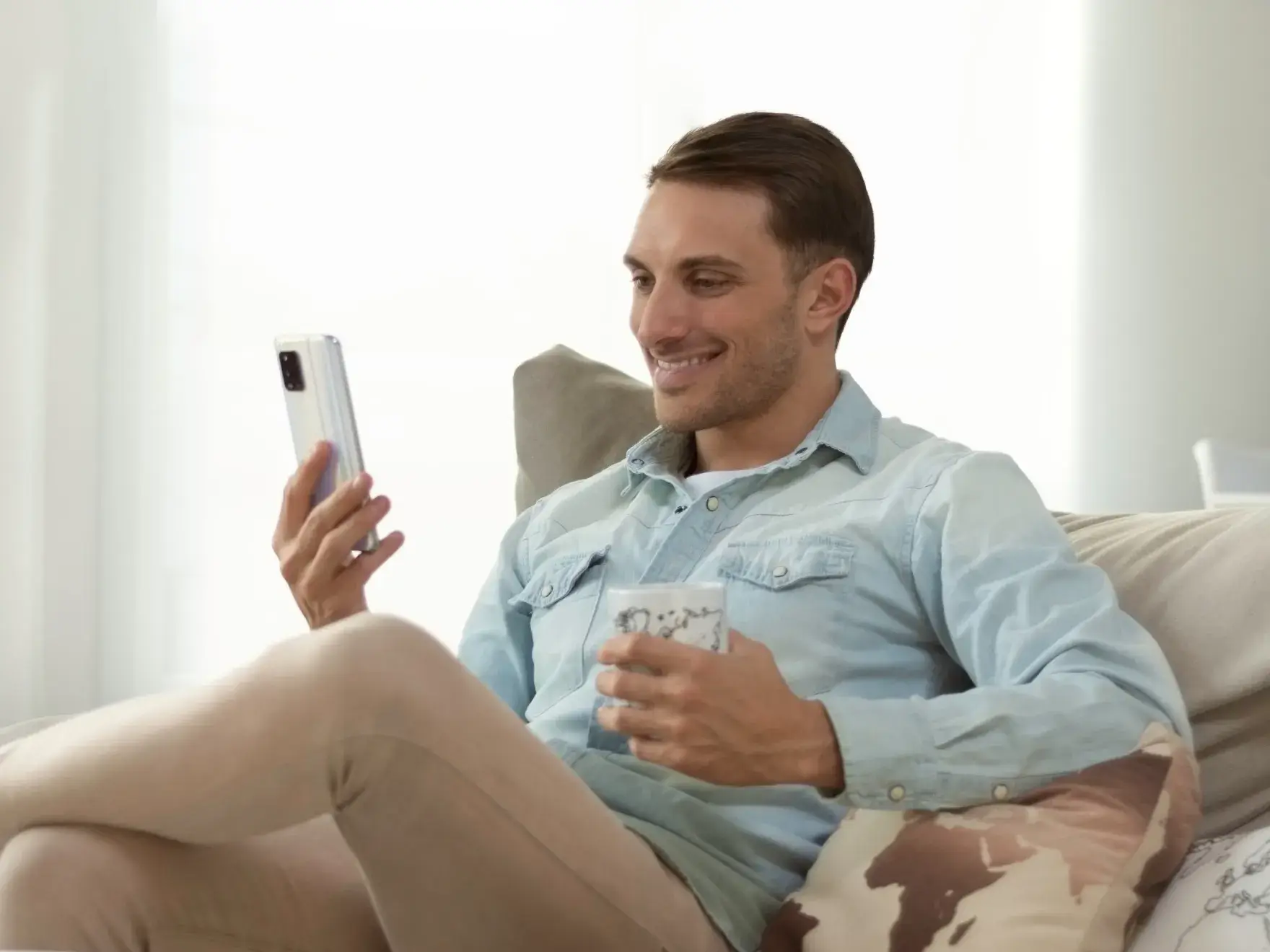 Man sitting on a couch holding a cup and looking at his smartphone with a smile.