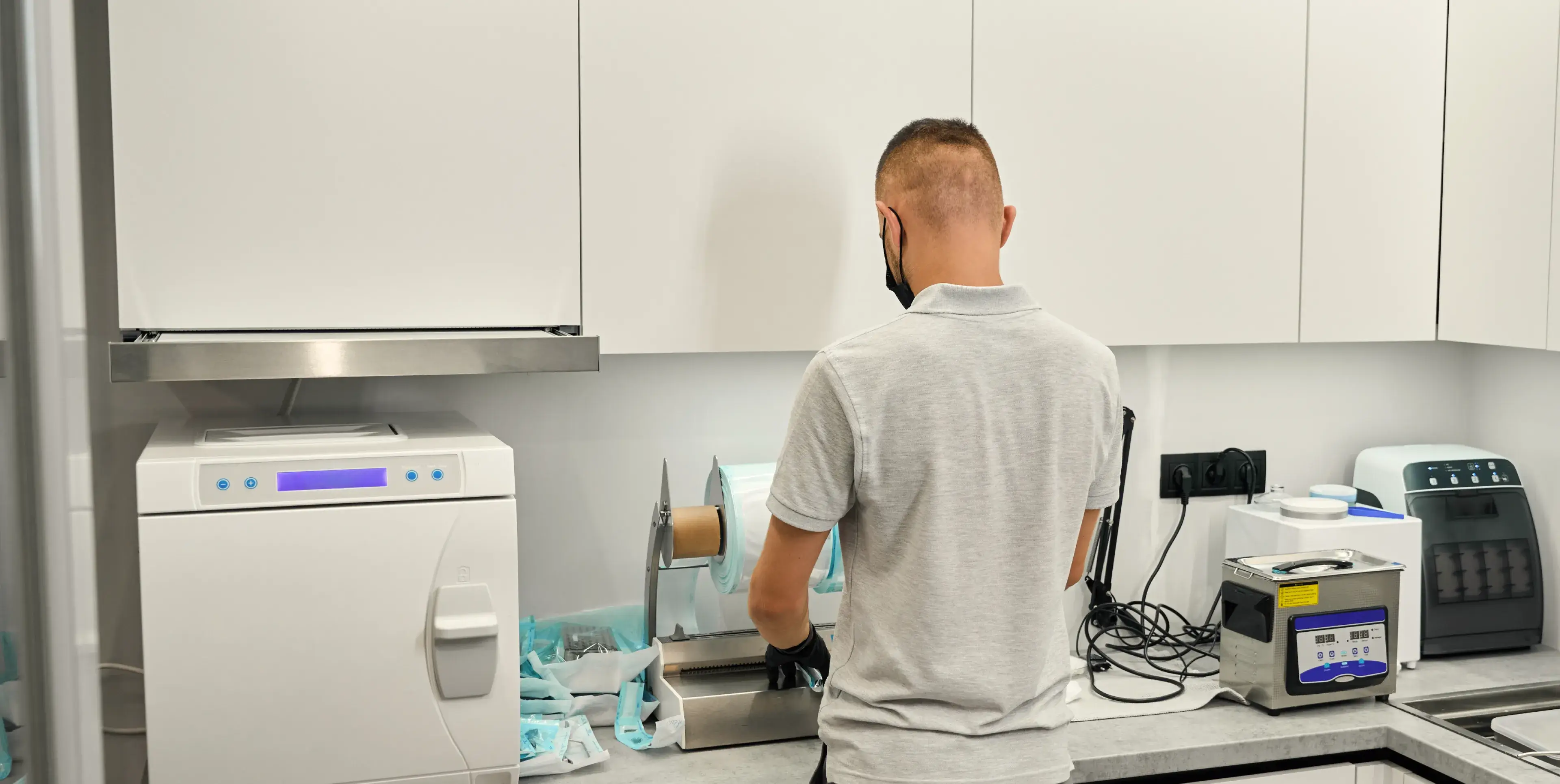 Person in a gray shirt and black gloves preparing sterilization pouches in a clean dental sterilization room.
