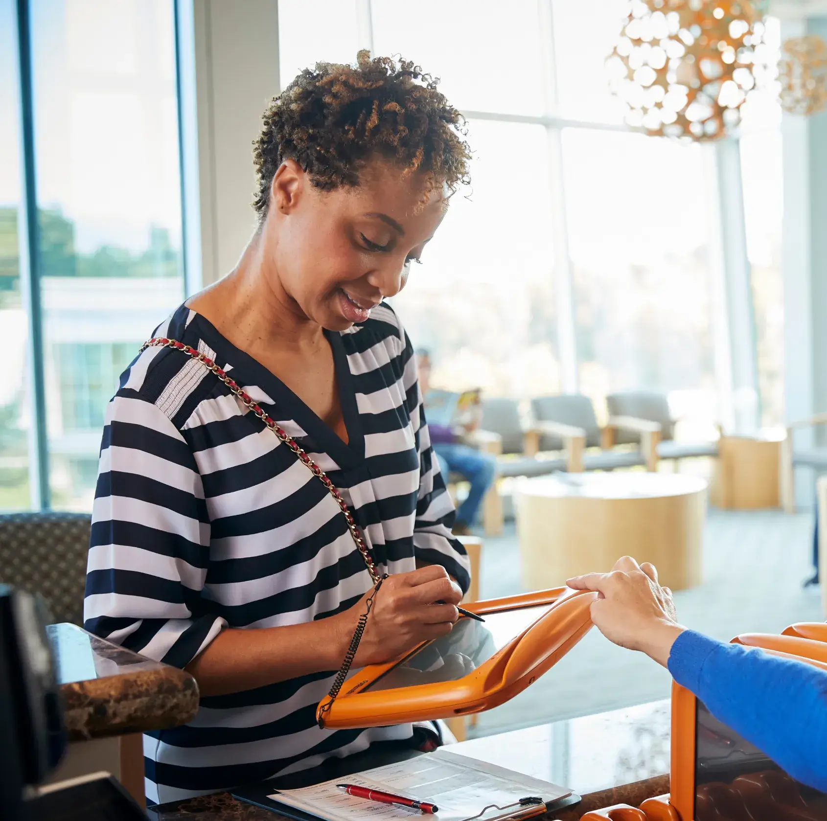 Smiling woman signing a digital tablet at a reception desk inside a bright waiting area.