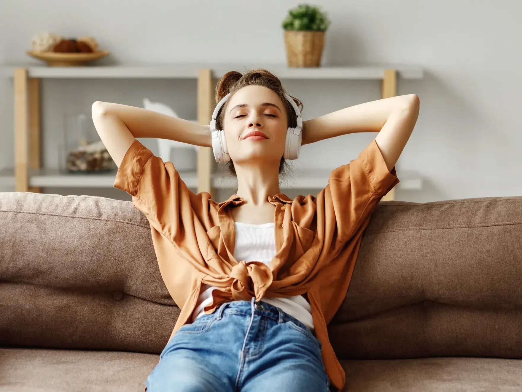 Young woman wearing white headphones relaxing on a brown couch with eyes closed and hands behind her head.