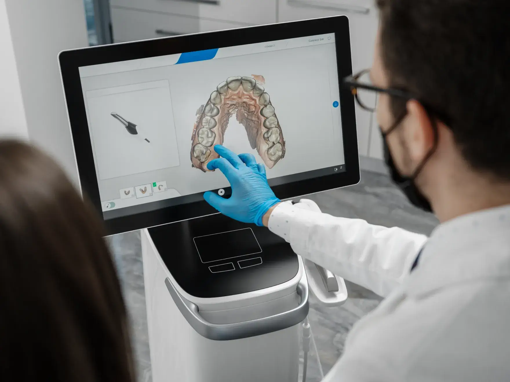 A dentist wearing blue gloves and a mask points at a 3D digital model of teeth on a screen in a dental office.