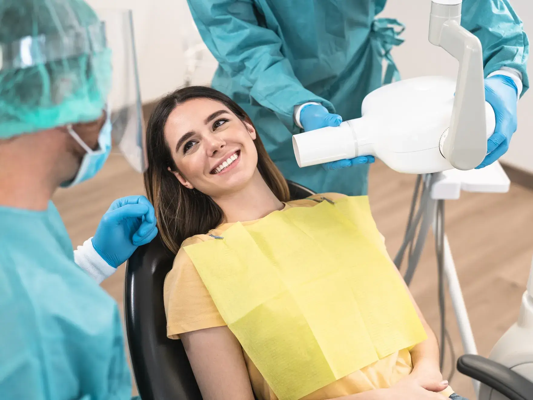 Smiling woman in a dental chair wearing a yellow bib, with two dental professionals in protective gear preparing dental equipment.
