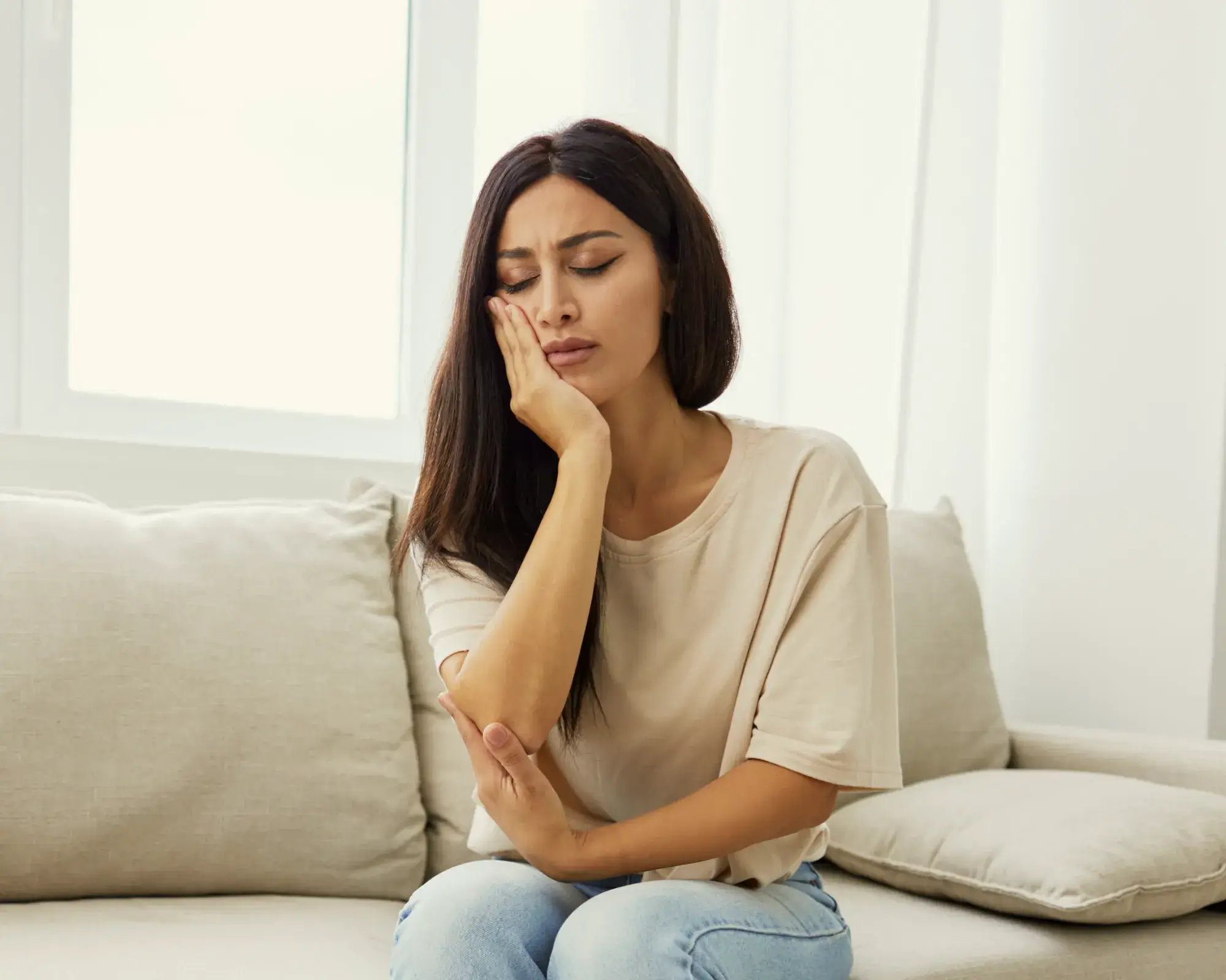 Woman sitting on a beige couch, holding her cheek with a pained expression, suggesting toothache or facial pain.