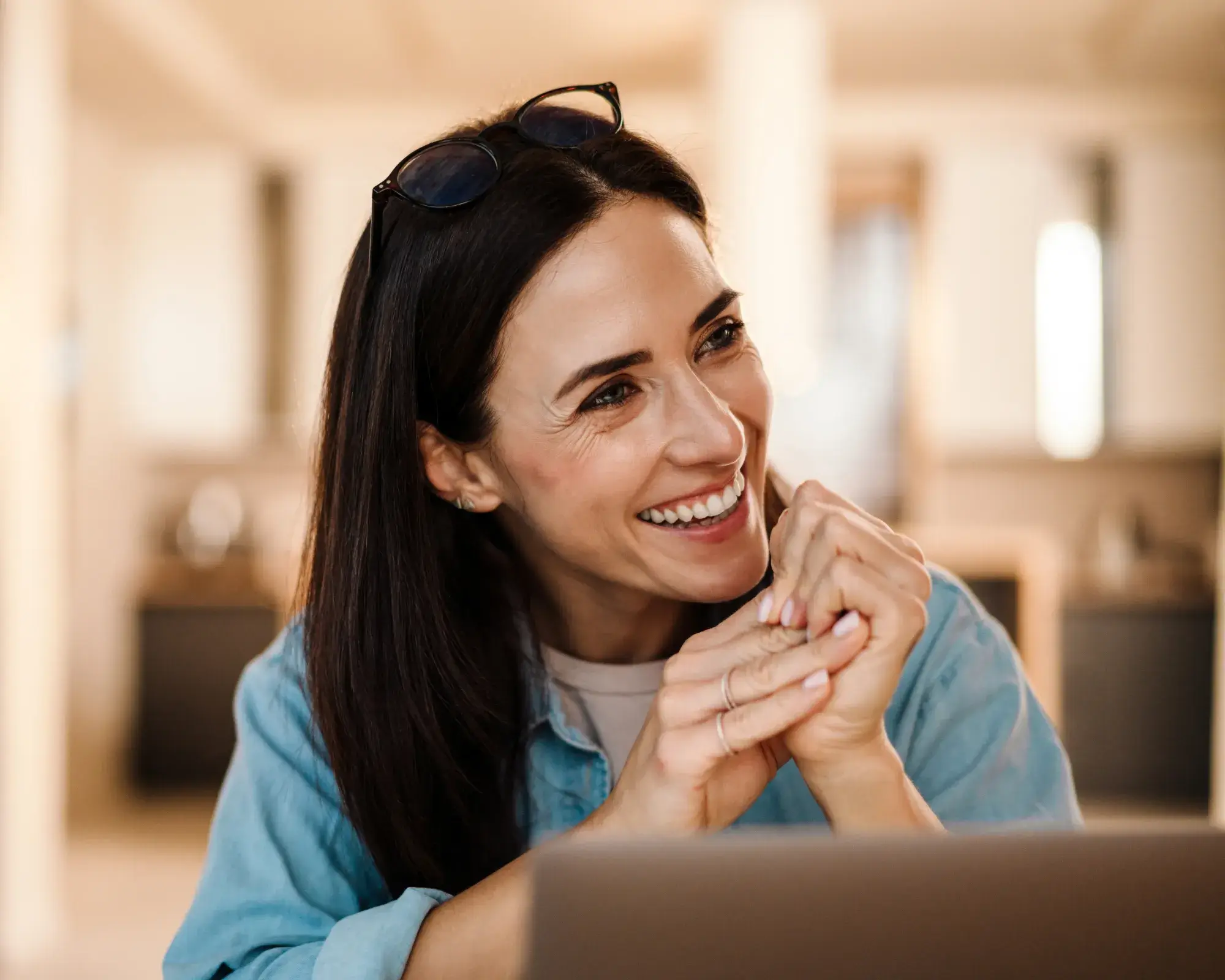 Smiling woman with long dark hair and glasses on her head, clasping her hands near a laptop.