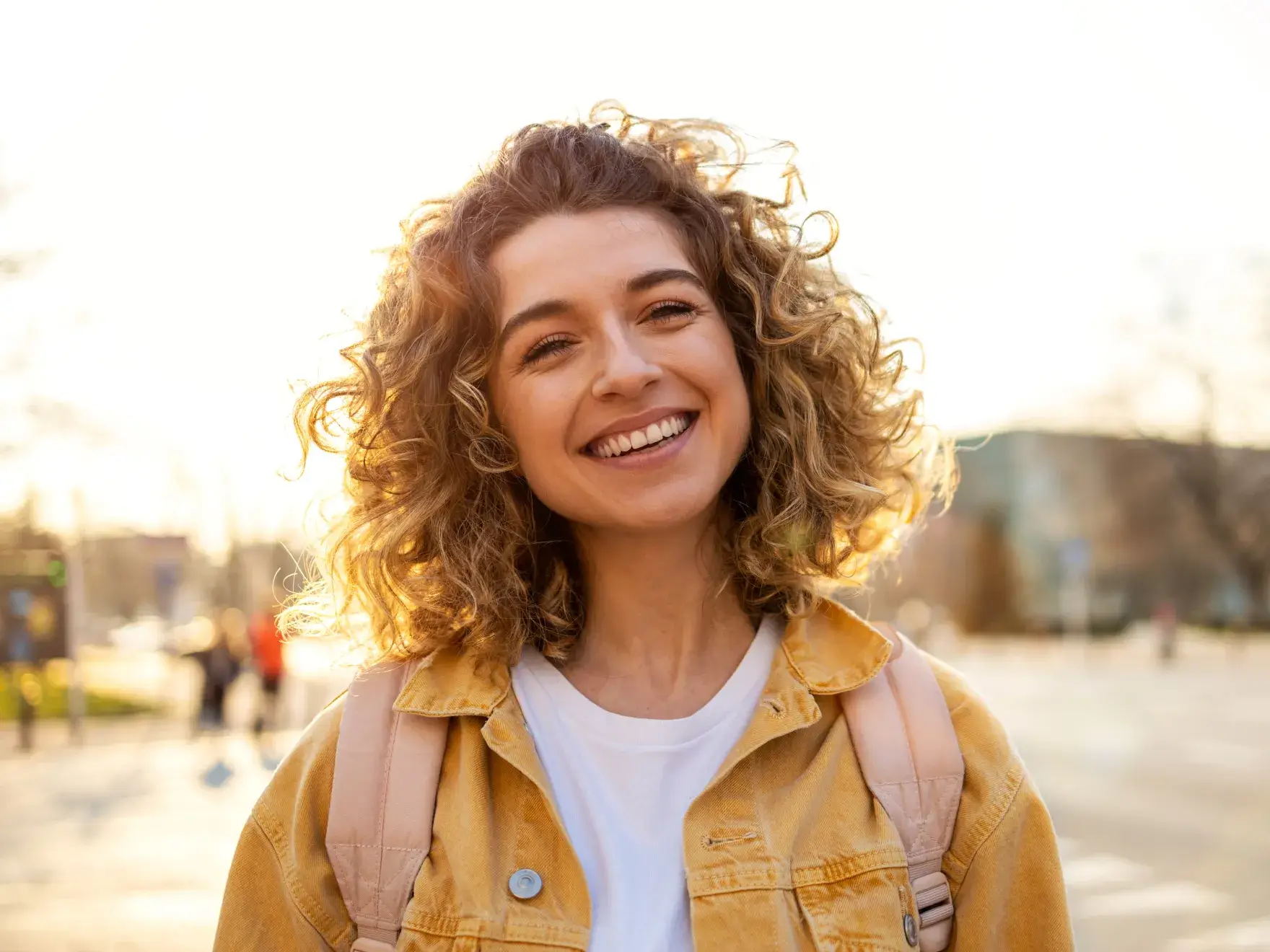Smiling young woman with curly hair wearing a yellow jacket and backpack outdoors at sunset.