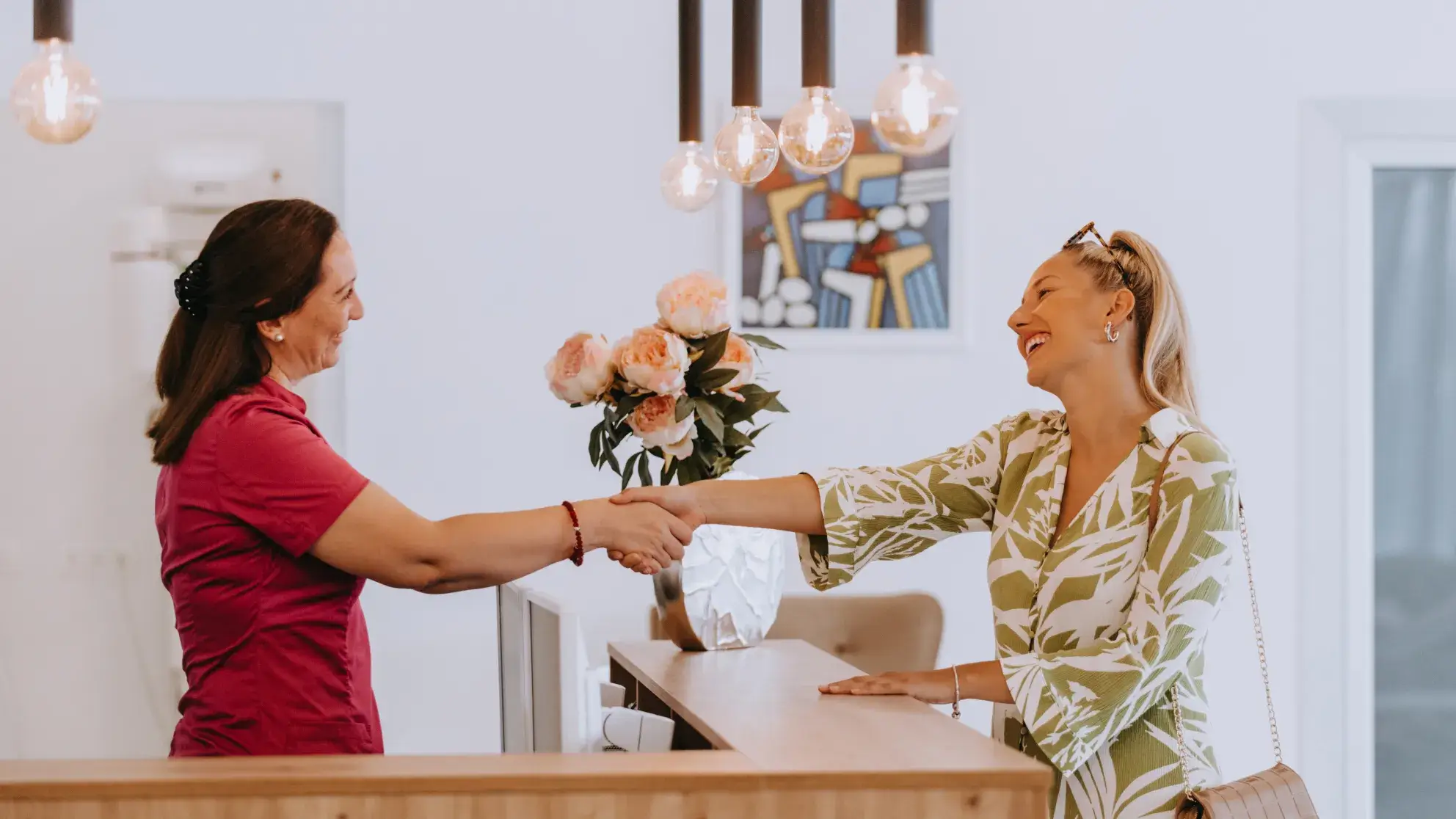 Two smiling women shaking hands across a reception desk with a vase of flowers in the background.