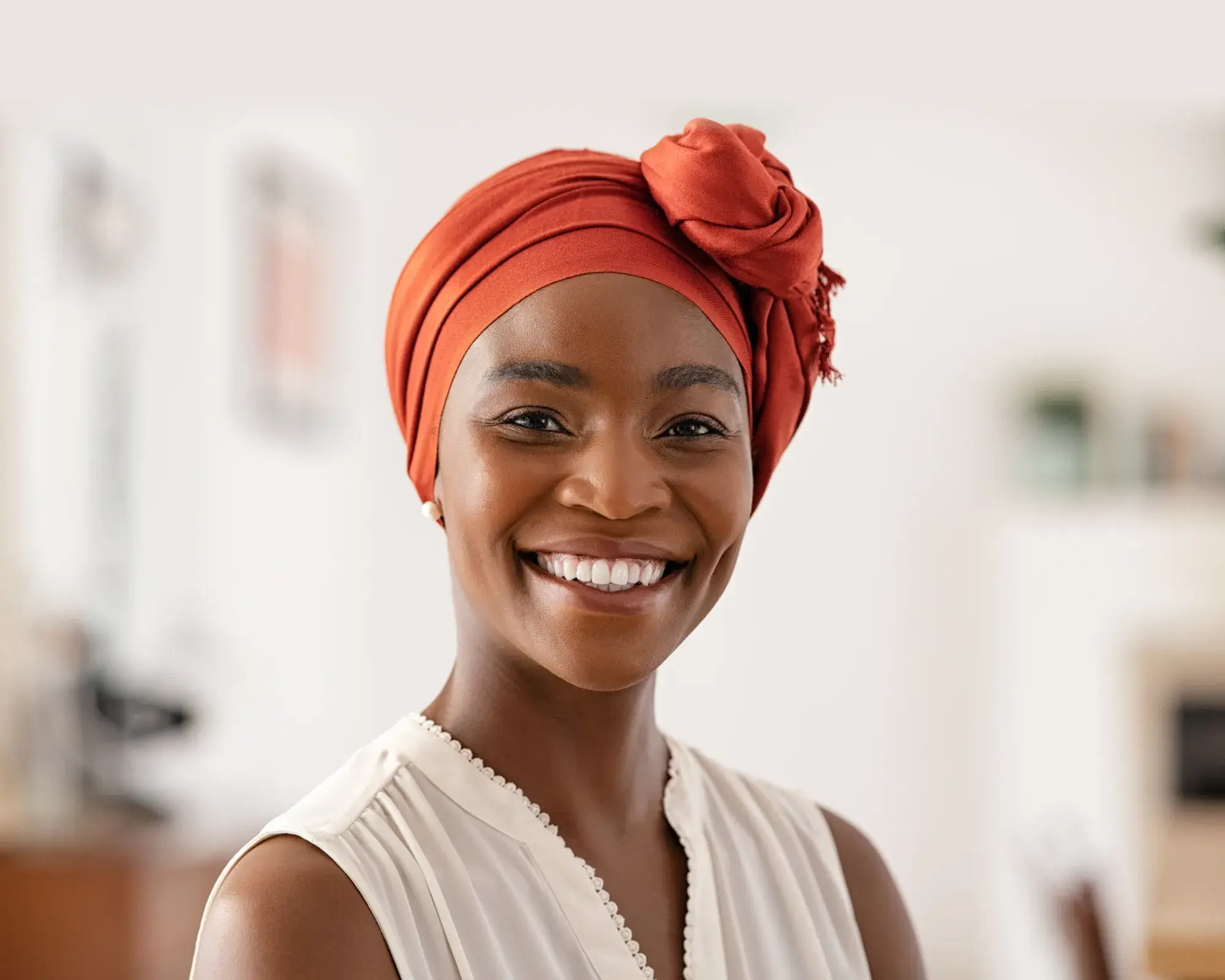 Smiling woman wearing an orange headwrap and white sleeveless top in a bright room.