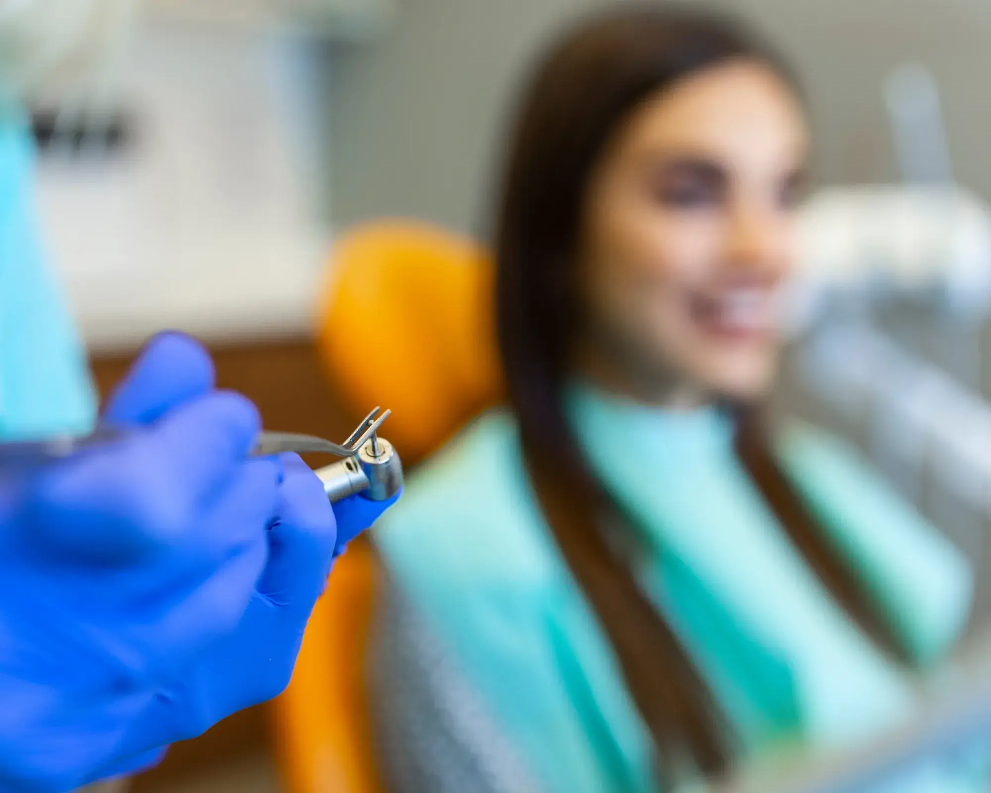 Dentist's gloved hand holding a dental drill near a smiling woman in a dental chair.