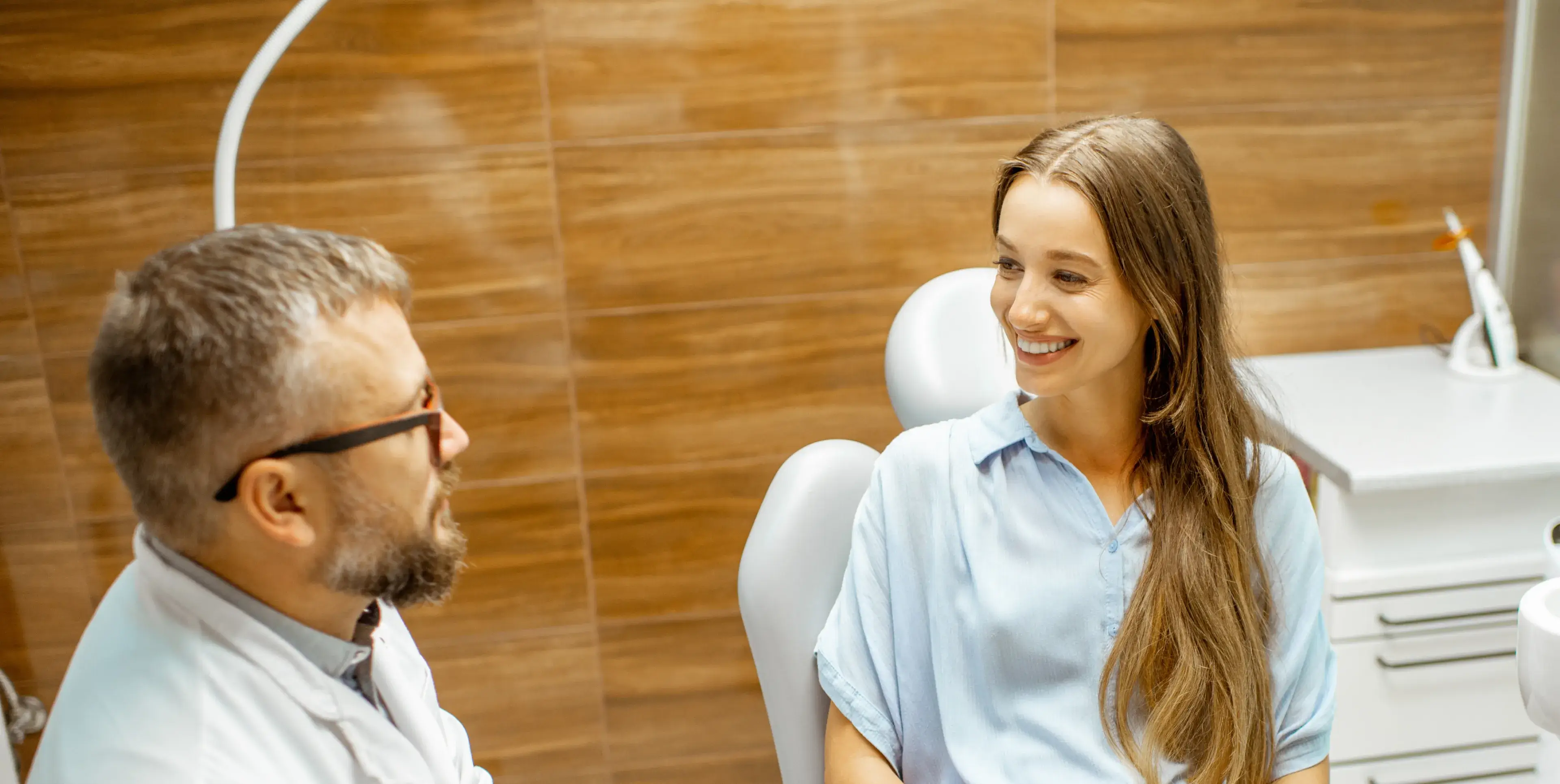 Male dentist with glasses talking to a smiling female patient seated in a dental chair.