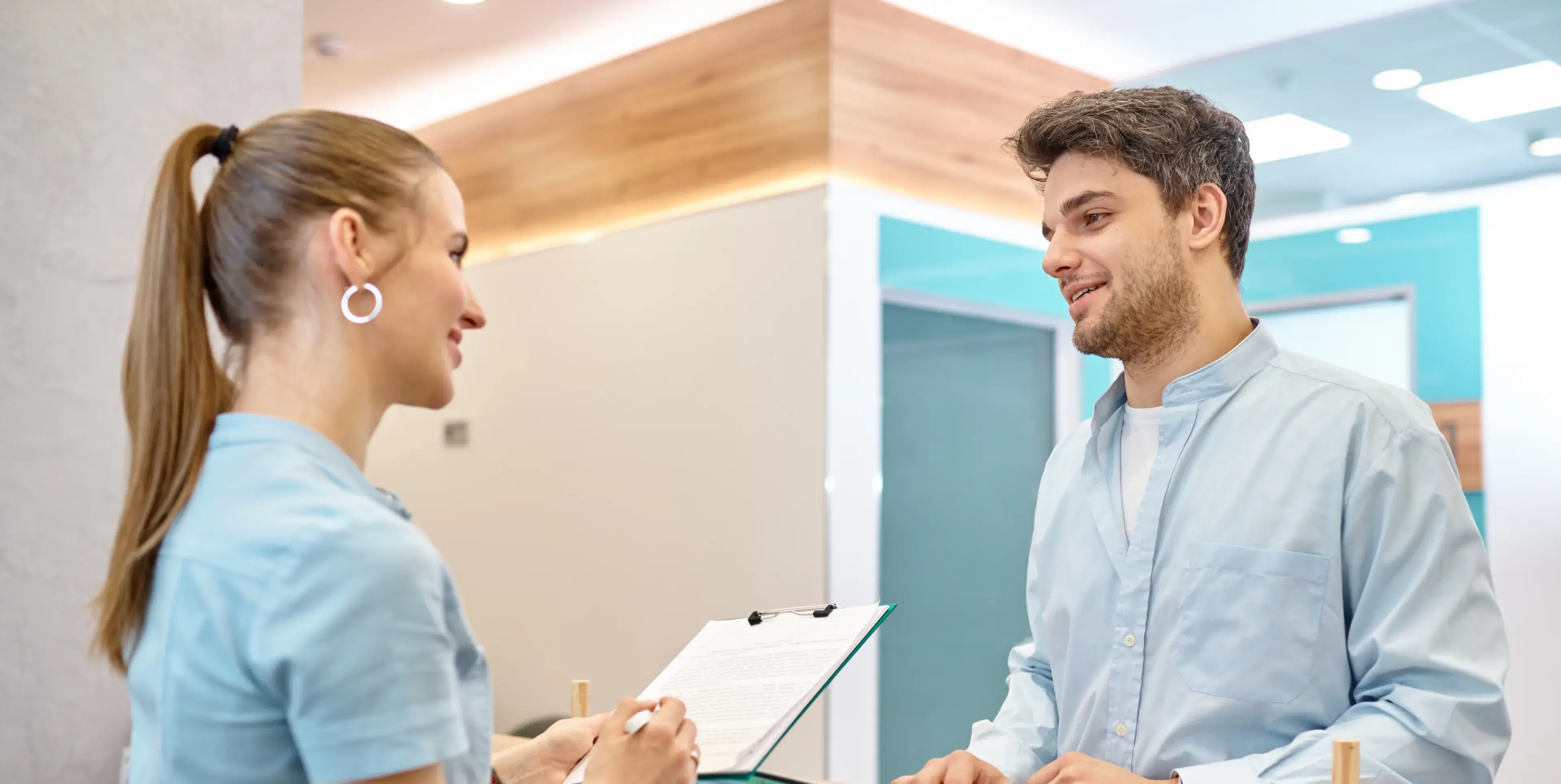 A woman holding a clipboard converses with a smiling man in a light blue shirt in a modern office setting.