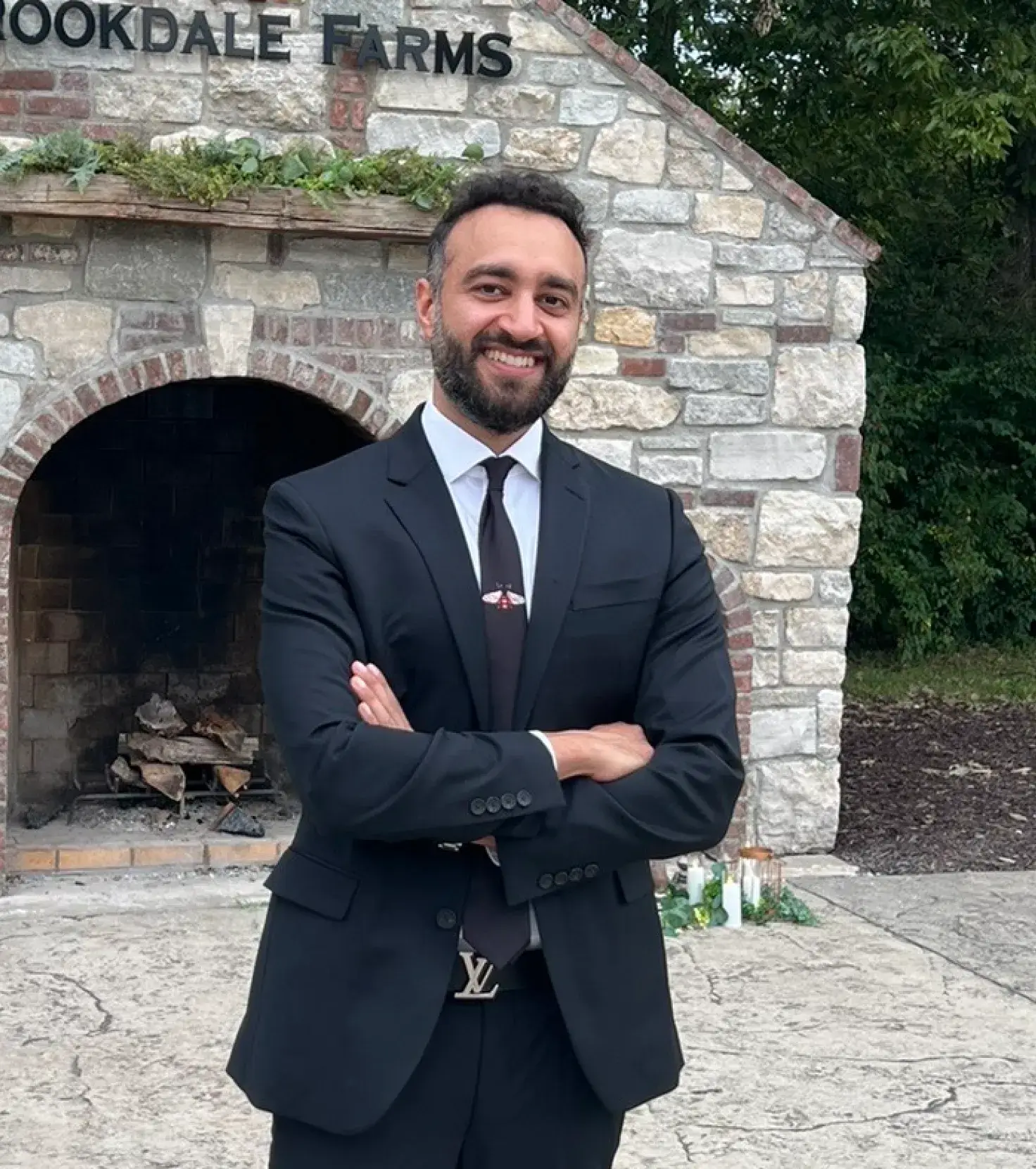 Man in a black suit and tie standing outdoors with arms crossed, smiling, in front of a stone fireplace with greenery on top and trees in the background.