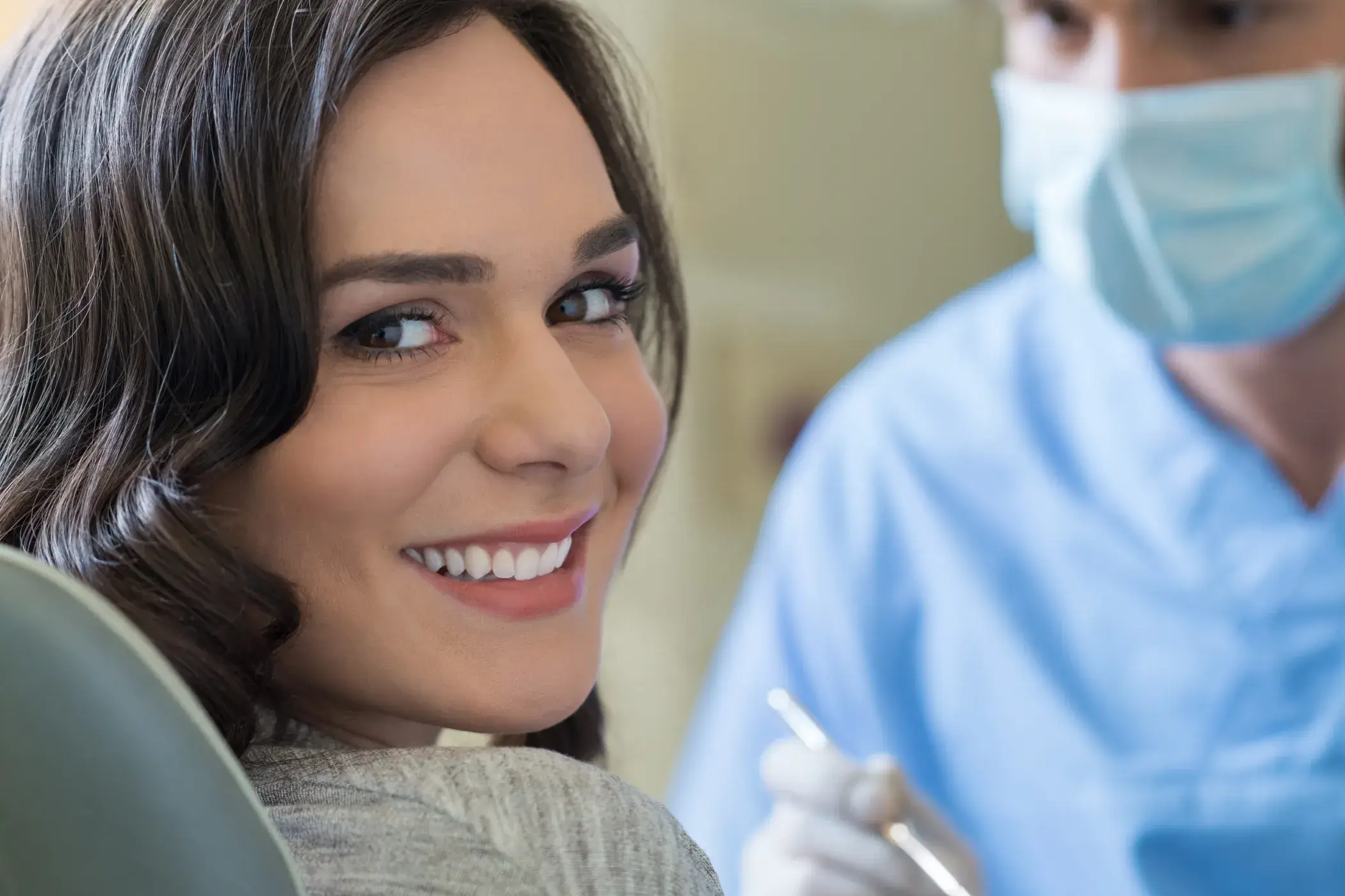 Smiling woman looking over her shoulder in a dental chair with a masked dentist holding tools in the background.