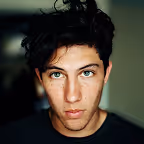 Young man with dark curly hair and freckles looking directly at the camera against a blurred background.