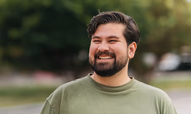 Homme souriant vêtu d'un t-shirt vert clair, avec une barbe et des cheveux foncés, en extérieur avec un arrière-plan flou d'arbres.