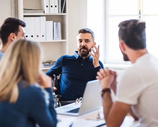 Homme en fauteuil roulant parlant à un groupe de collègues assis autour d'une table avec un ordinateur portable.
