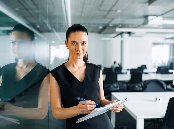 Femme debout dans un bureau moderne, tenant un stylo et un carnet, regardant vers l'avant avec confiance.