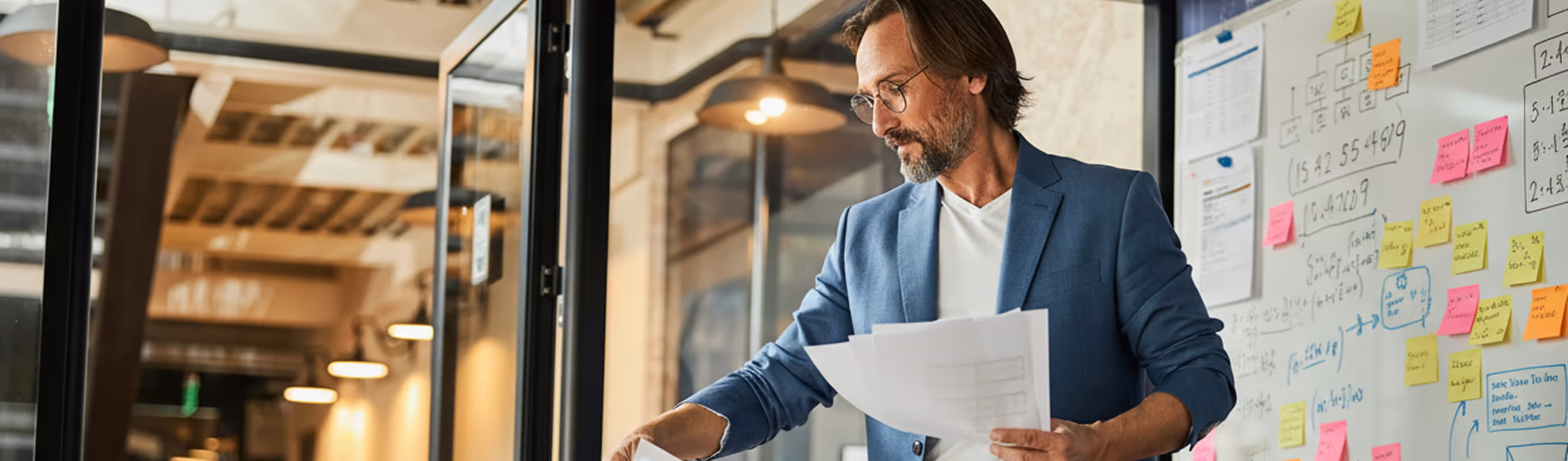 Homme d'affaires avec des lunettes et une veste bleue examinant des papiers dans un bureau moderne avec un tableau blanc couvert de notes colorées et d'écrits.
