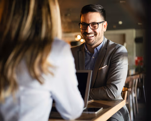 Un homme souriant en costume gris et lunettes discute avec une femme aux cheveux longs dans un café, avec un ordinateur portable sur la table.
