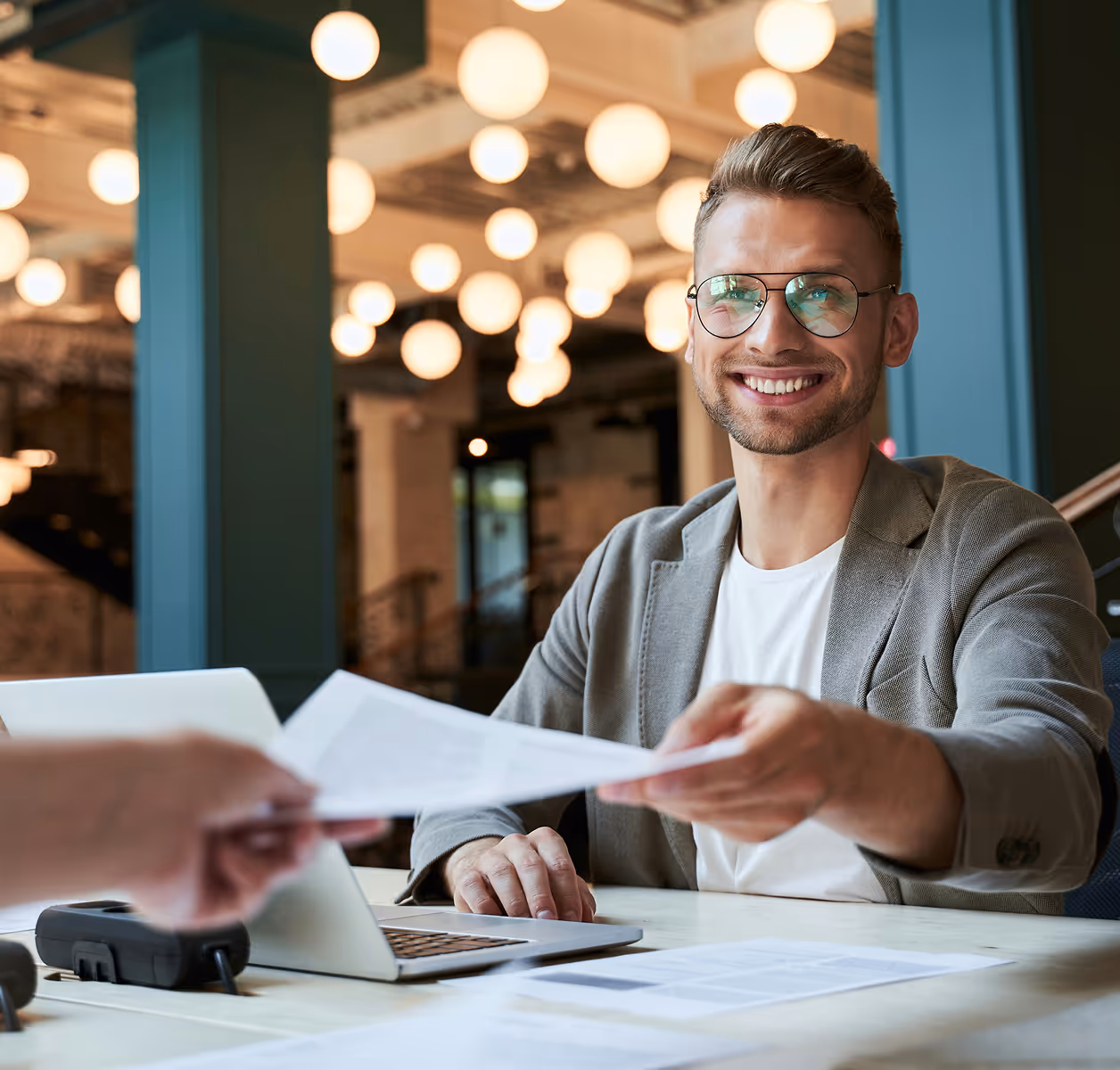 Jeune homme souriant portant des lunettes, assis à une table avec un ordinateur portable, tendant des documents à une autre personne dans un bureau moderne éclairé par des lampes suspendues.