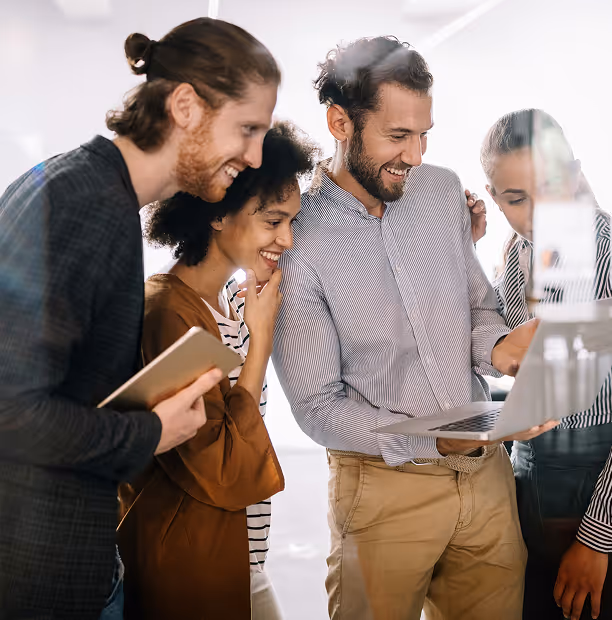 Quatre jeunes professionnels souriants regardant un ordinateur portable ensemble dans un environnement de bureau lumineux.