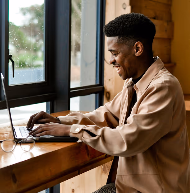 Un homme souriant tape sur un ordinateur portable près d'une fenêtre dans un café en bois.
