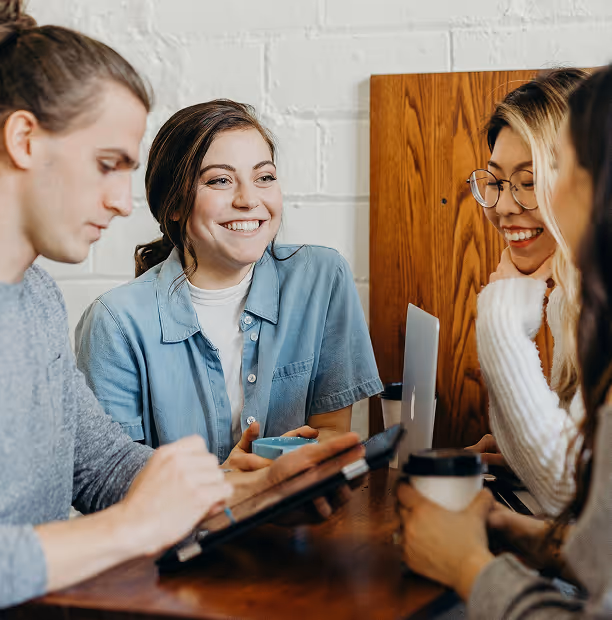 Quatre jeunes adultes souriants discutant et travaillant ensemble autour d'une table avec un ordinateur portable et des boissons.