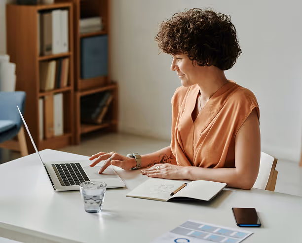 Jeune femme souriante travaillant sur un ordinateur portable à une table blanche avec un carnet ouvert et un verre d'eau.
