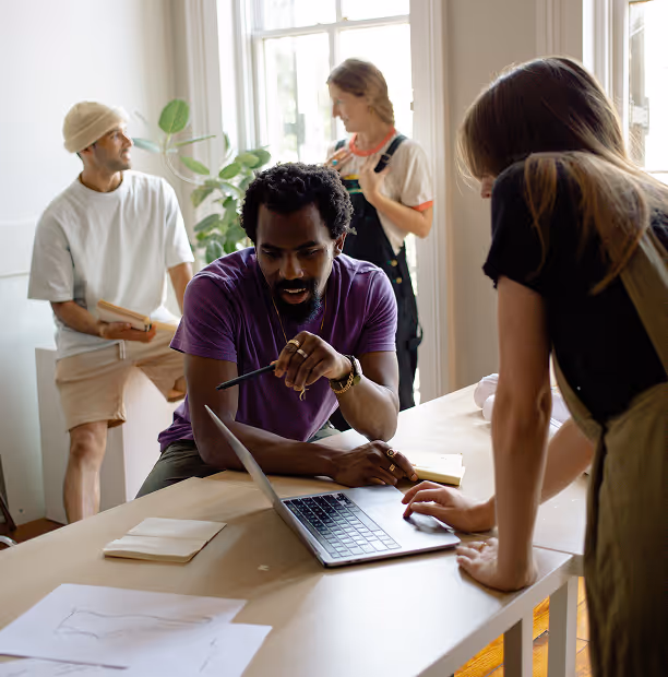 Quatre jeunes adultes collaborent autour d'un ordinateur portable dans une salle lumineuse, deux discutant près de la table tandis que deux autres sont à l'arrière.
