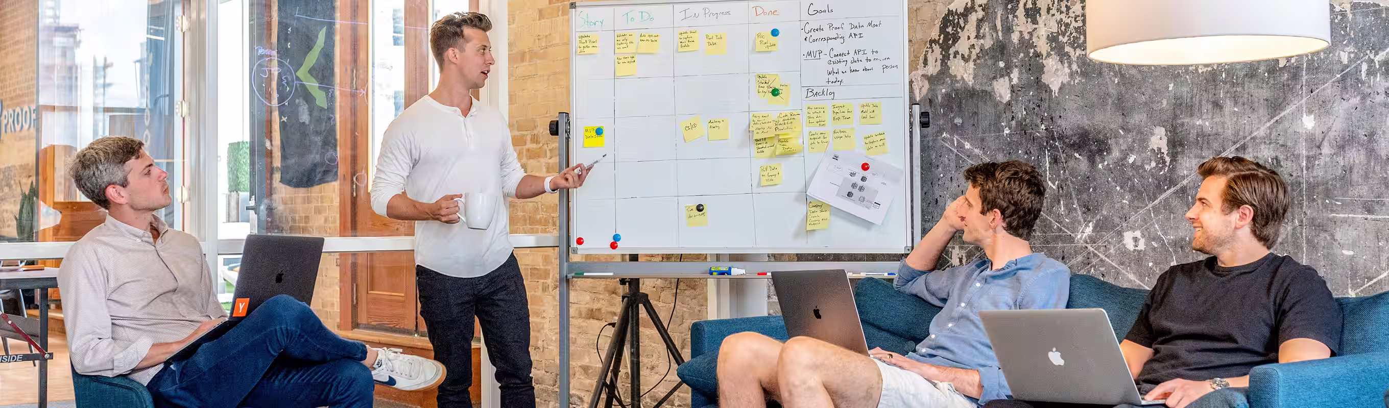 Quatre hommes en réunion dans une salle moderne, l'un debout présente devant un tableau blanc avec des notes adhésives, les autres sont assis avec des ordinateurs portables.