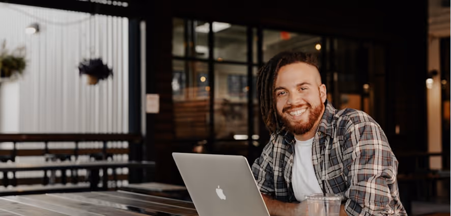 Un homme souriant avec une chemise à carreaux utilise un ordinateur portable Mac dans un café moderne.