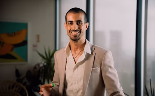 Un homme souriant portant un costume beige avec une chemise blanche, debout dans une pièce avec une plante et une grande fenêtre en arrière-plan.