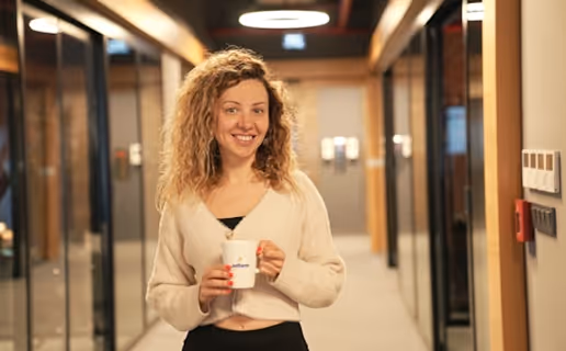 Jeune femme souriante tenant une tasse blanche dans un couloir de bureau moderne.