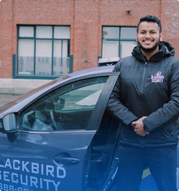 Smiling security guard standing beside a Blackbird Security vehicle with open door, in front of a brick building.